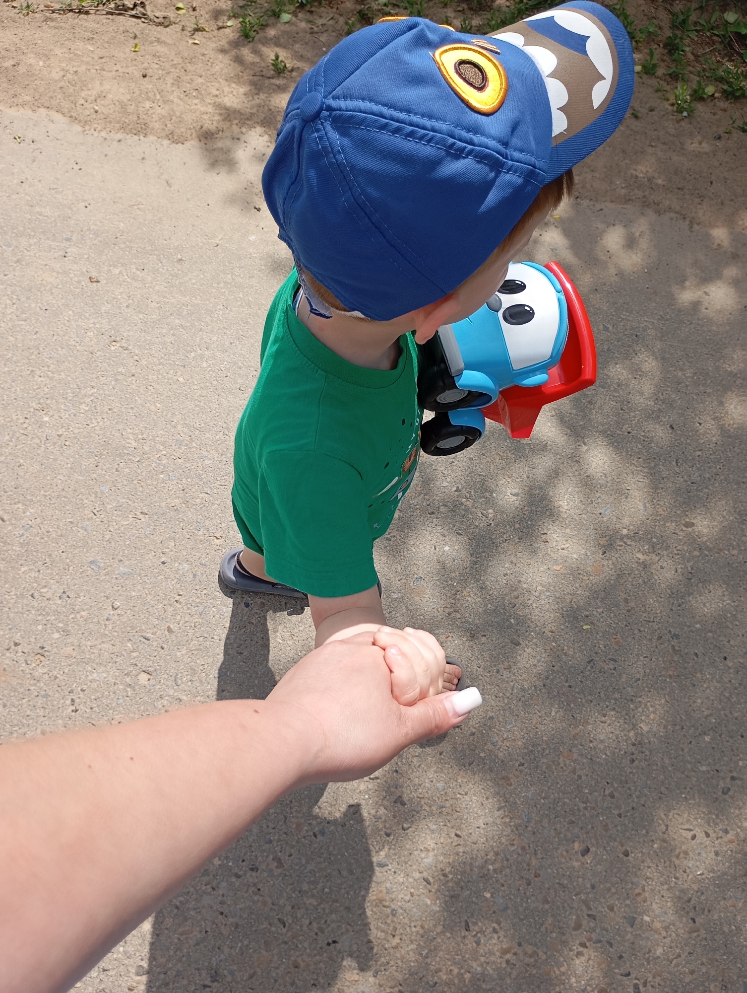 A high-angle shot depicts a toddler wearing a blue baseball cap adorned with cartoonish eye and mouth designs. He sports a green shirt and gray shoes. The child is holding a blue and red toy truck. An adult's hand is visible in the lower-left, seemingly holding the toddler's hand. The scene appears to be outdoors on a paved surface.