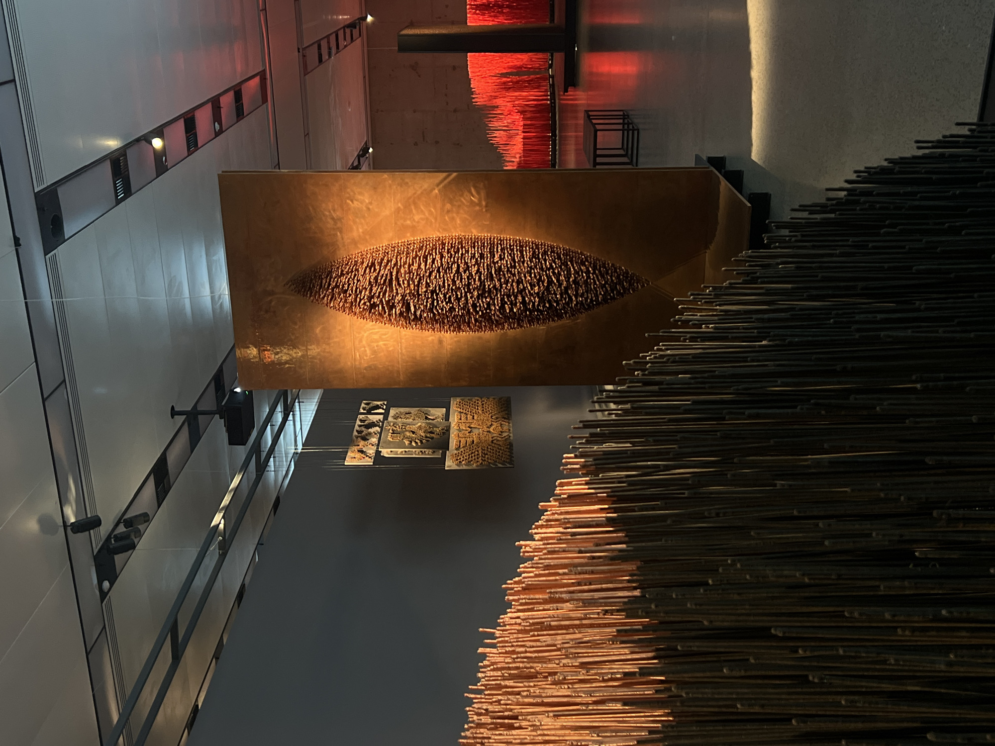 The image depicts the interior of the Zaryadye Park in Moscow, Russia. Various art installations are featured prominently. One installation consists of a large, horizontally oriented, oblong shape filled with small, dark brown elements mounted on a bronze-colored rectangular panel. Another installation involves numerous slender rods, possibly metallic, stacked densely. The bottom section of this installation is colored in copper-like tones, transitioning upwards to darker, near-black tones. Several smaller, rectangular artworks are placed nearby. The building's interior reveals architectural details, including modern walls, ceiling lights, and metal framework. Overhead, a separate installation involving suspended, glowing red strands is visible.