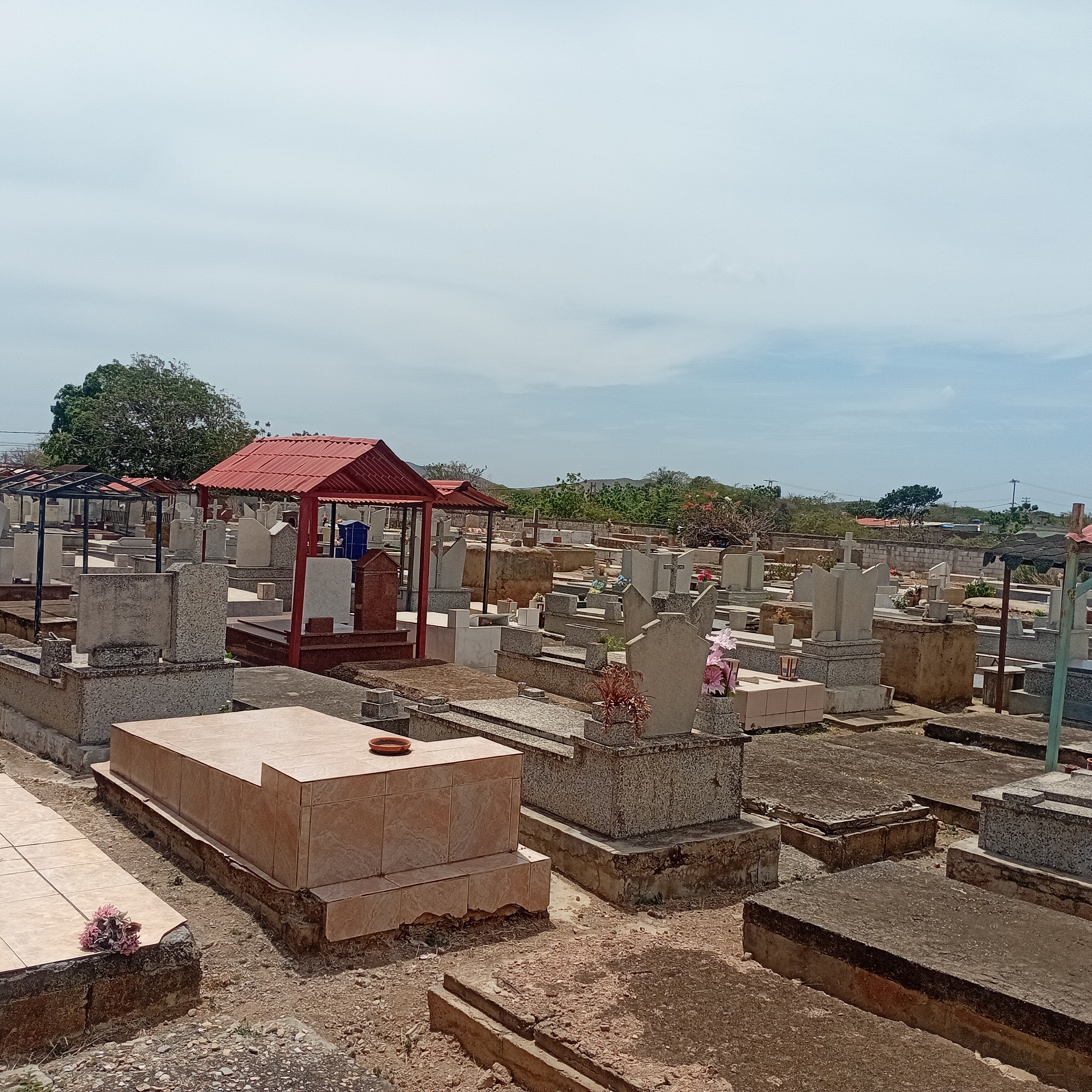 The image shows a cemetery located in an unknown city. The scene contains numerous graves and tombstones of varying sizes and styles. Some graves are covered with tiles, while others are made of stone or concrete. Some graves have red canopies. The ground surrounding the graves is covered in gravel. In the background, there are trees and some buildings. The sky is blue with some clouds.