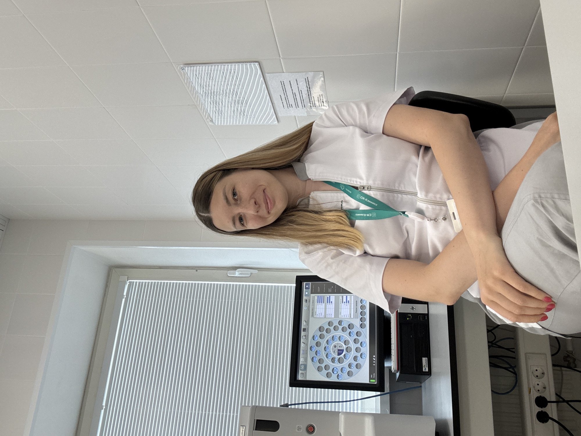 A woman wearing a white lab coat and grey pants is seated in a chair in what appears to be a laboratory or clinical setting. She is looking towards the camera with a slight smile. She has long blonde hair. In front of her is a desk with a computer monitor displaying a circular interface with multiple circular sections, a keyboard, and a computer tower with "HP" and "SYSMEX" branding. To the left of the monitor is a piece of laboratory equipment with a red button. The background consists of white tiled walls and a window with horizontal blinds. There are also some electrical outlets and cables visible on the wall and under the desk. A green lanyard with a badge is hanging around the woman's neck. The scene is indoors and appears to be well-lit. There are no visible indicators that this location is in Moscow, Russia.