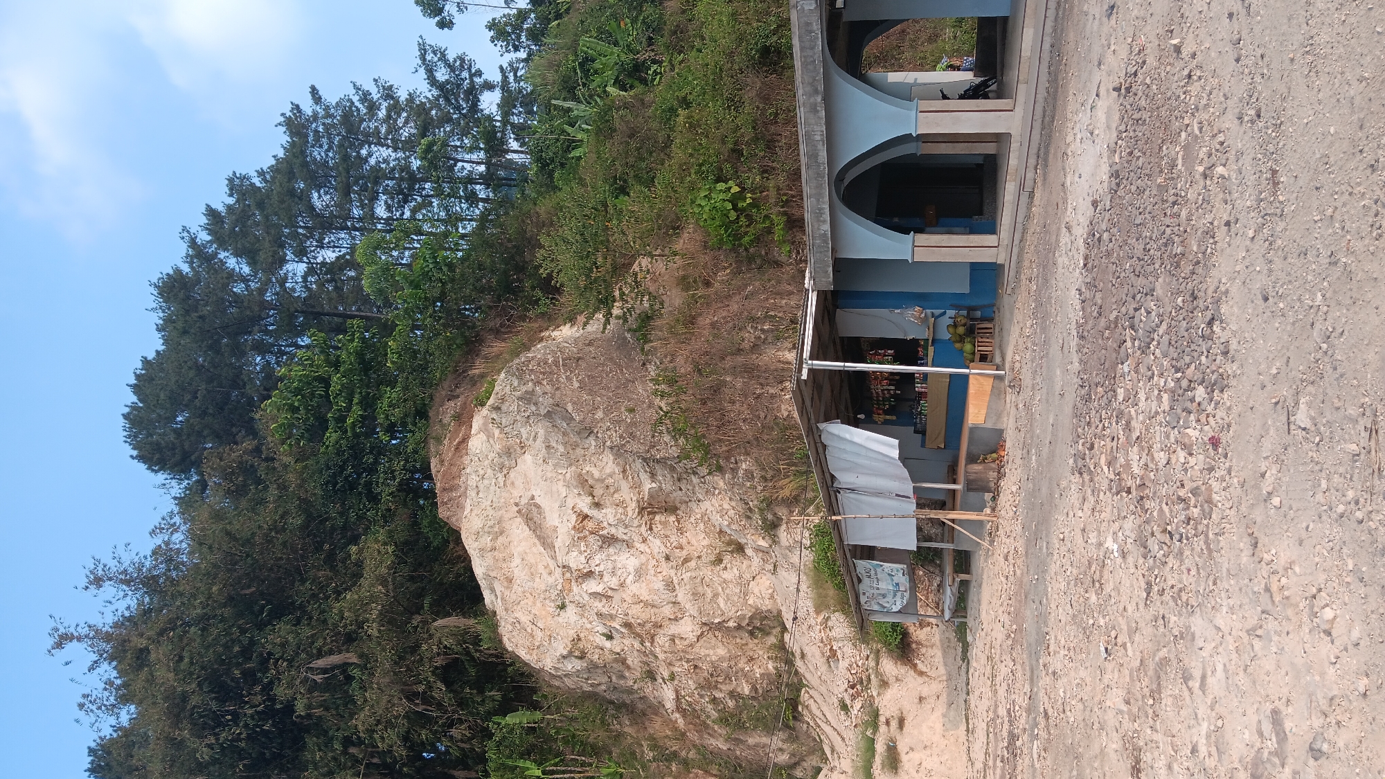 The image depicts an outdoor scene with a small roadside stall or shop in the foreground, set against a backdrop of a rocky hillside covered in lush vegetation. The stall has a white canopy and a blue-painted structure with arched openings. Various items are displayed on shelves within the stall, and a crate of coconuts is visible to the right. The ground in front of the stall is rough and unpaved, covered in gravel and dirt. Behind the stall, a large, light-colored rock formation dominates the hillside, with dense green trees and foliage growing on and around it. The sky above is a clear blue with a few wispy clouds. The scene appears to be located in a rural or semi-rural area. Based on the provided context, this location is identified as being in the city of Mekargalih, Indonesia. No people are visible in the image.