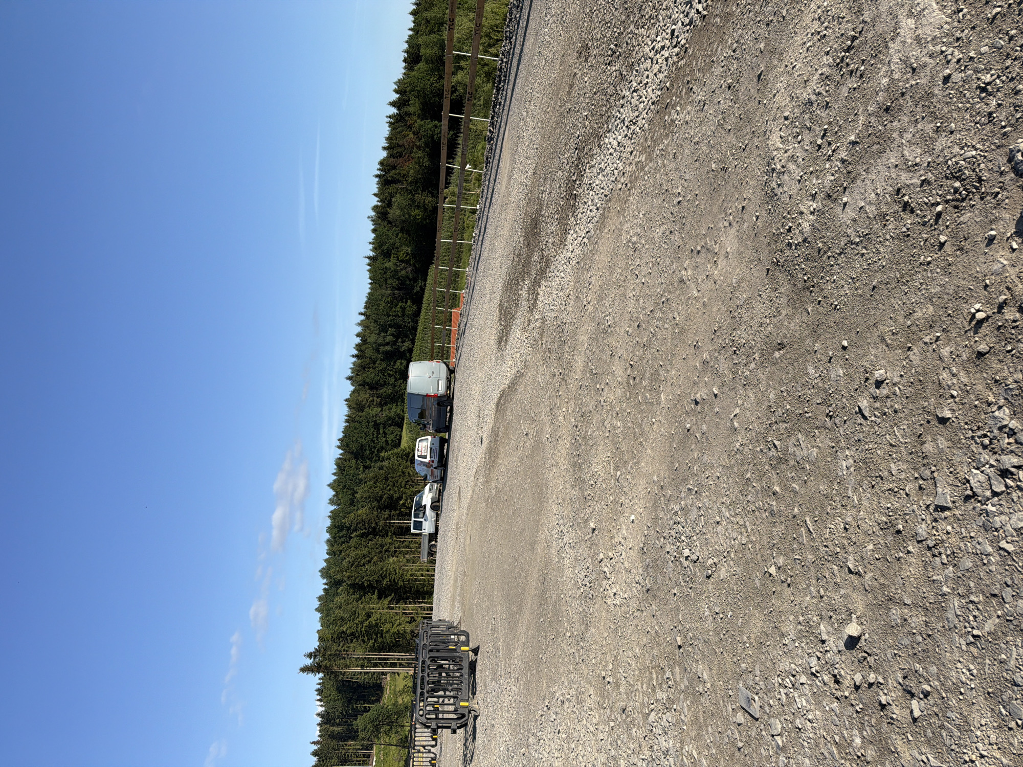 A clear, sunny day in Stróża, Poland, with a vast expanse of blue sky dominating the upper portion of the image. Below the sky, a dense, dark green coniferous forest stretches horizontally across the horizon, its trees tightly packed. In the foreground, a rough, gravel-covered area occupies the majority of the frame, appearing as a parking lot or construction site. Several vehicles are parked on the gravel. To the left, a black, segmented barrier stands, seemingly for crowd control or temporary fencing. Further into the scene, a white flatbed truck is parked, followed by two white vans, and a dark grey van or SUV. A wooden fence with several white posts and visible signage runs along the edge of the gravel area, bordering the treeline. The overall impression is one of an outdoor, possibly rural, location with evidence of recent activity or preparation.