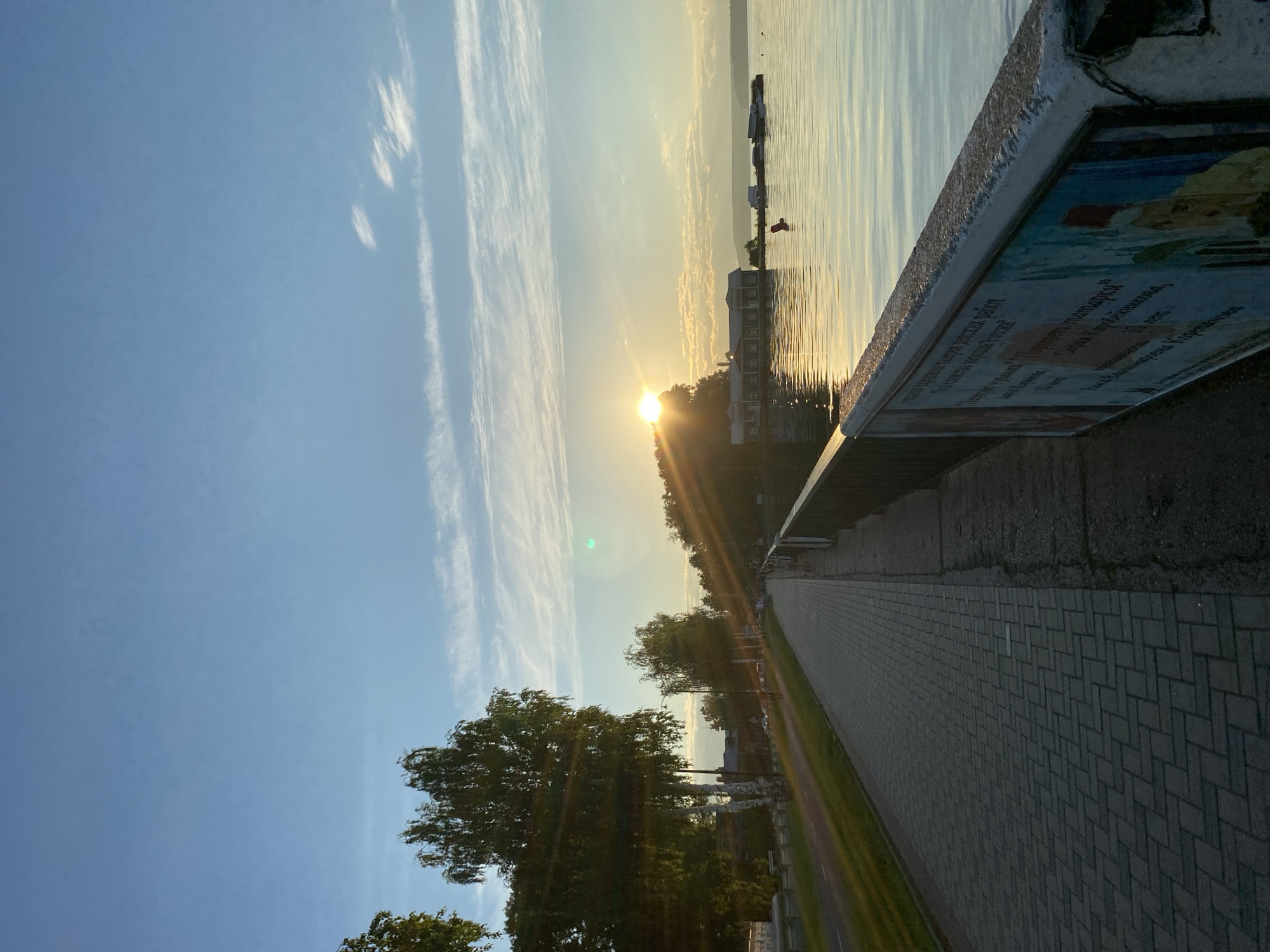 The image depicts a scenic waterfront promenade in Votkinsk, Russia, likely during a sunrise or sunset. The sun, partially obscured by trees on a distant embankment, casts a warm glow across the scene. Long, thin clouds streak across the clear blue sky.
On the left side of the image, a paved pedestrian walkway stretches into the distance, bordered by a grassy area and trees. A road runs parallel to the walkway, with some cars visible. The trees closest to the viewer are birch trees with characteristic white bark.
On the right, a railing separates the walkway from a calm body of water, reflecting the sky and sunlight. In the distance across the water, there is a line of buildings with dark roofs, possibly a resort or residential area. Several boats are moored near the shore. A bright red buoy is visible in the water. The railing itself has a concrete structure, and on its visible side, there is a faded advertisement or mural with text and images. The overall atmosphere is tranquil and serene.