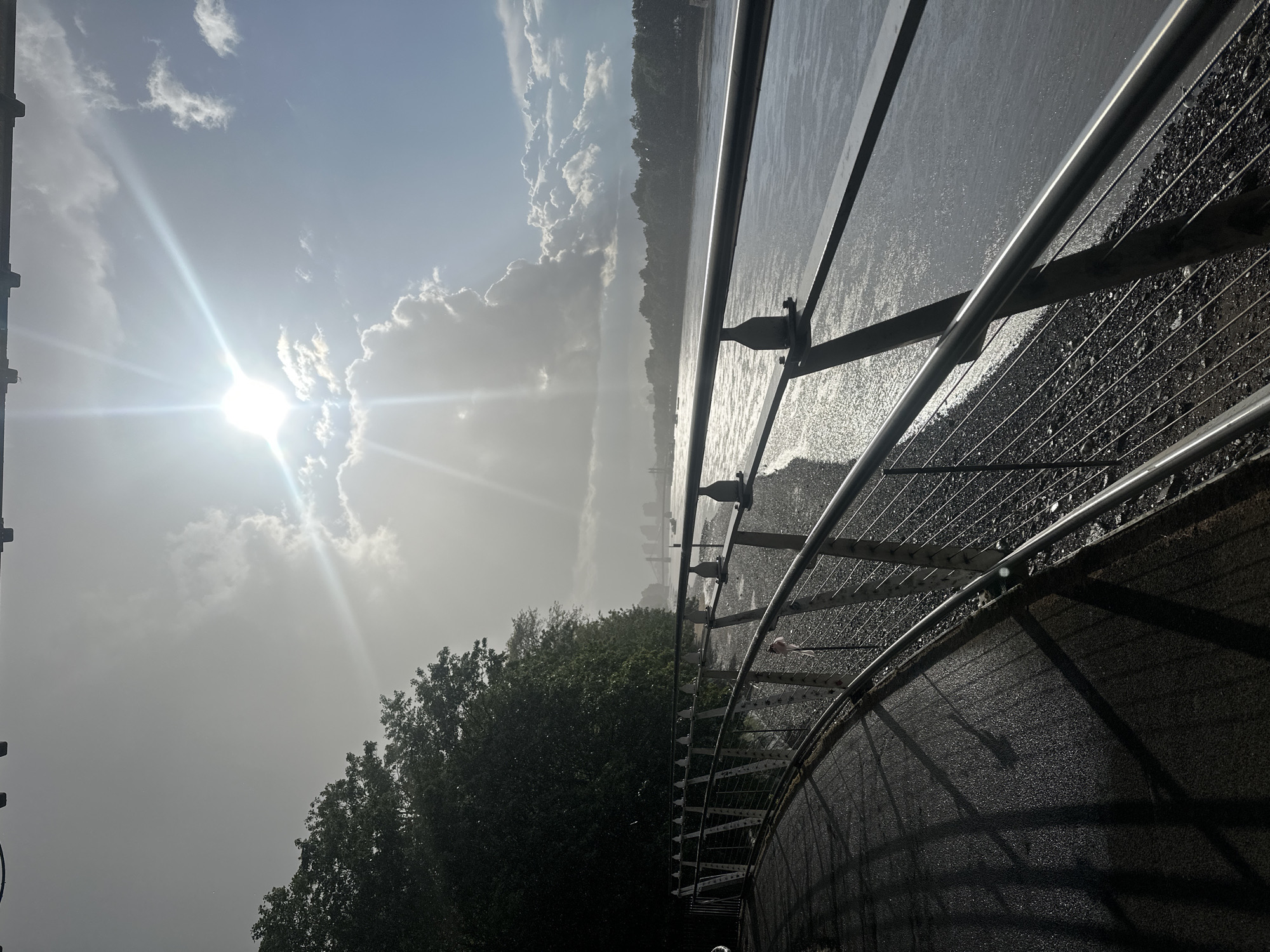 The image captures a view overlooking the River Thames in London, United Kingdom, on a bright but cloudy day. A metal railing runs along the right side of the frame, suggesting a walkway or bridge. Beyond the railing, the river reflects the sunlight, creating a shimmering effect. Trees line the riverbank in the midground, and in the distance, a blurred skyline of buildings can be seen. The sky is filled with clouds, with the sun shining brightly, creating a strong glare. The image appears to be taken from a slightly elevated position.