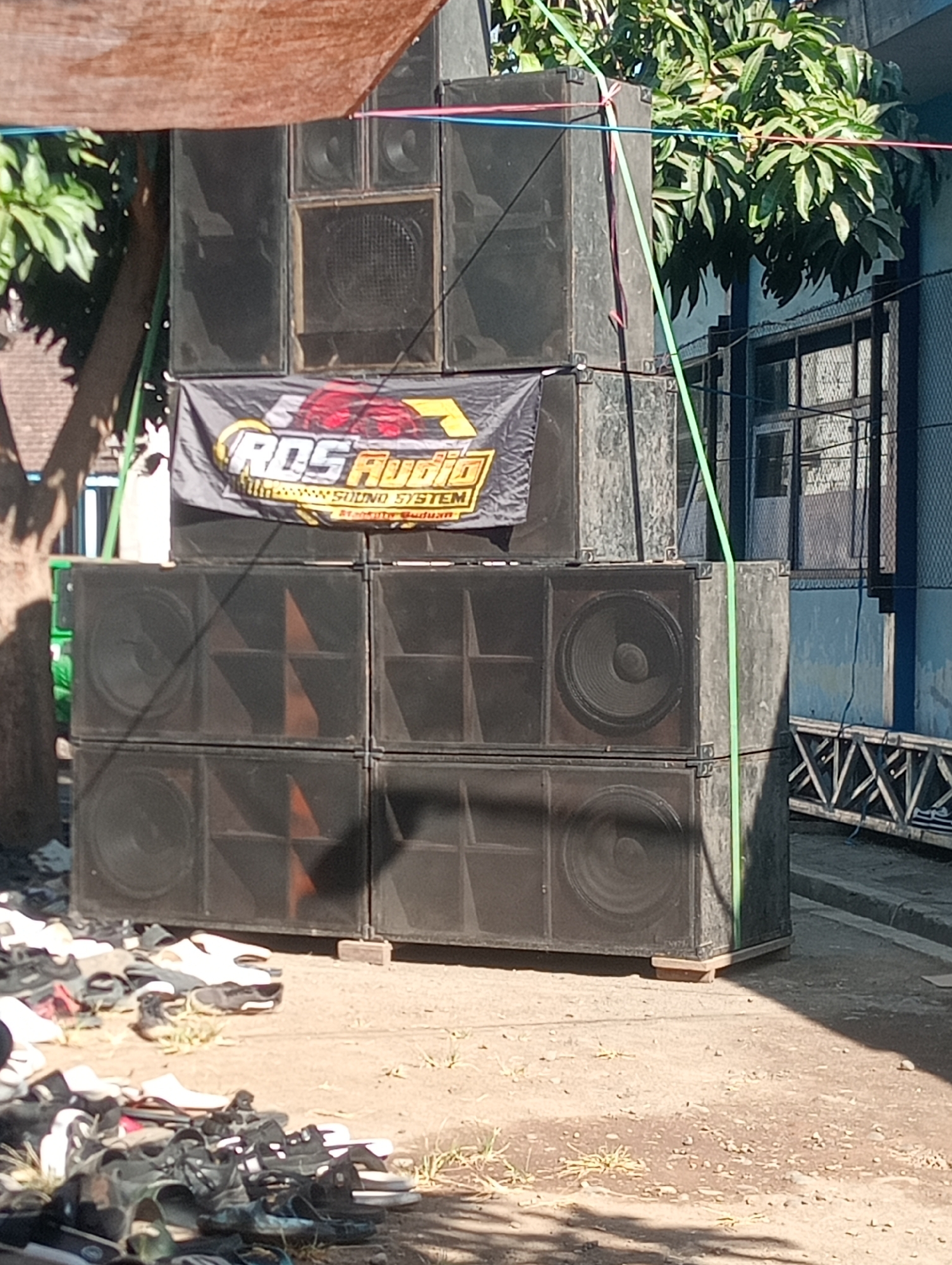 The image depicts an outdoor scene in Buduan, Indonesia, featuring a large stack of black audio speakers. A banner with the text "ROS Audio Sound System Buduan" is draped across the front of the speakers. The speakers are arranged in multiple tiers, with some units featuring large circular speaker cones. In the foreground, a collection of various footwear, including sandals and shoes, is scattered on the ground. The background shows a building with windows and a fence, and trees with lush green foliage. A wooden structure with metal criss-cross patterns is visible on the right side of the image. The scene appears to be set up for an outdoor event or gathering.