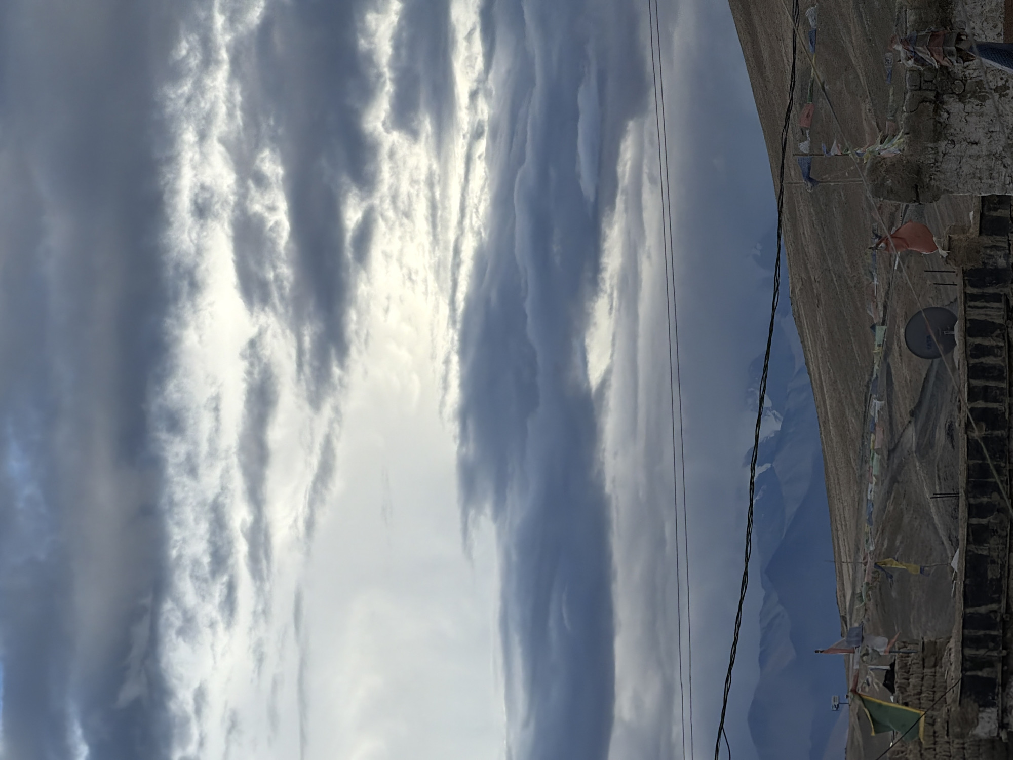 The image captures a wide shot of a mountainous landscape under a dramatic, cloudy sky. The sky is filled with layers of dark and light gray clouds, with patches of bright white light breaking through, suggesting an overcast or stormy weather condition. In the foreground, a series of power lines stretch horizontally across the frame. Below the power lines, the terrain is rugged and appears to be a hillside. Prayer flags in various colors are strung up and flutter in the wind, attached to posts or structures on the hillside. A satellite dish is visible on the left side of the frame, mounted on a wall. The lower portion of the image shows parts of what appears to be buildings or structures, made of rough stone or brick. In the far background, a range of majestic, snow-capped mountains rises against the horizon. The scene is likely set in a high-altitude, remote area. Given the presence of prayer flags and the mountainous terrain, this location could be in a region like Ladakh, India, which is consistent with Karzok, India.