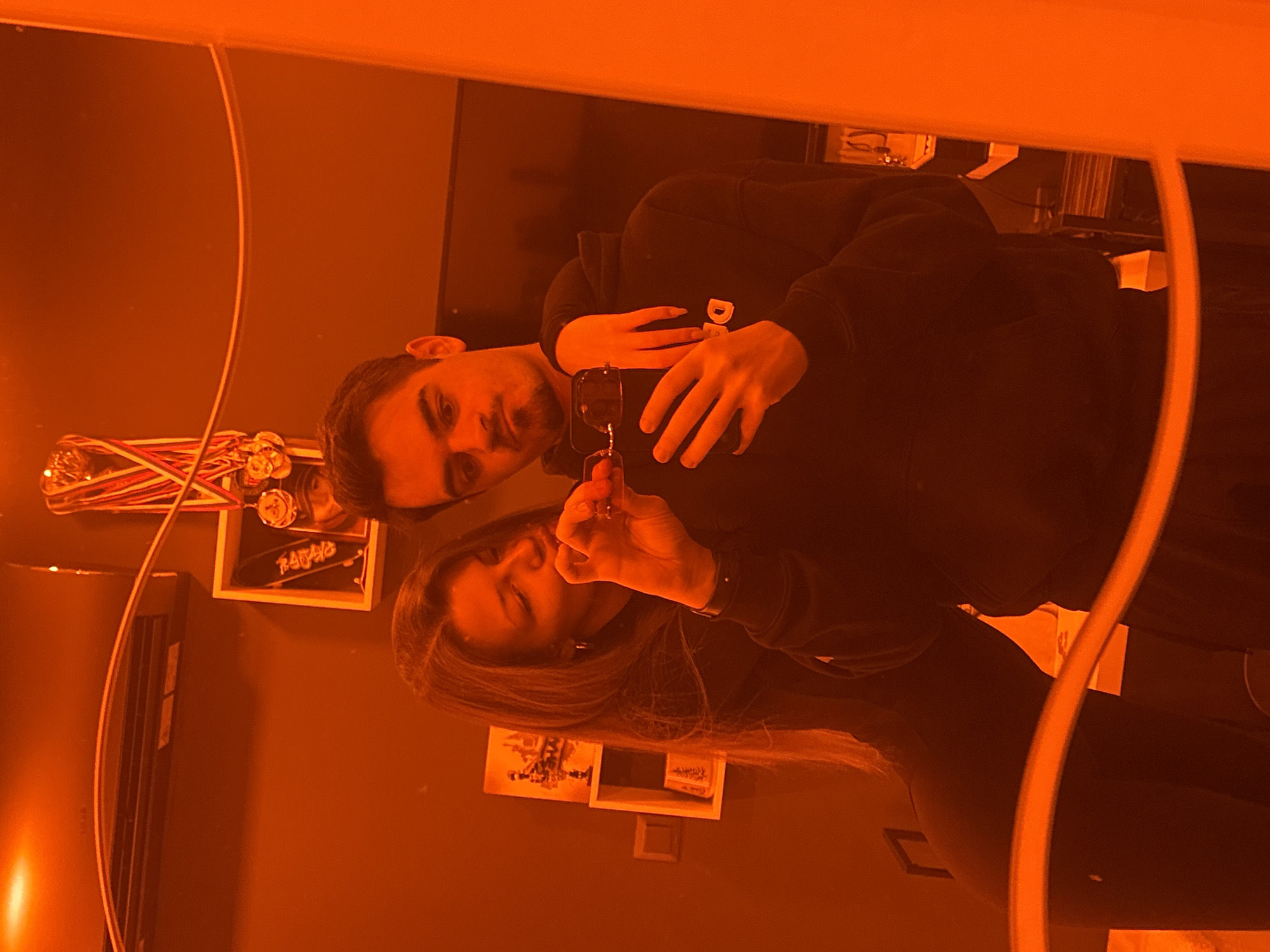 A man and a woman are reflected in a mirror. The man is holding a black smartphone and wearing a dark-colored hooded sweatshirt. The woman, also wearing dark clothing, has long brown hair.
Behind them are various objects mounted on a dark-colored wall, including medals, a framed picture with the word "RAFALO" written on it, a radiator, and other framed artwork. Other objects are positioned on a shelf visible in the background.
The lighting is strongly orange. There is no definitive information to confirm the location is Sofia, Bulgaria.