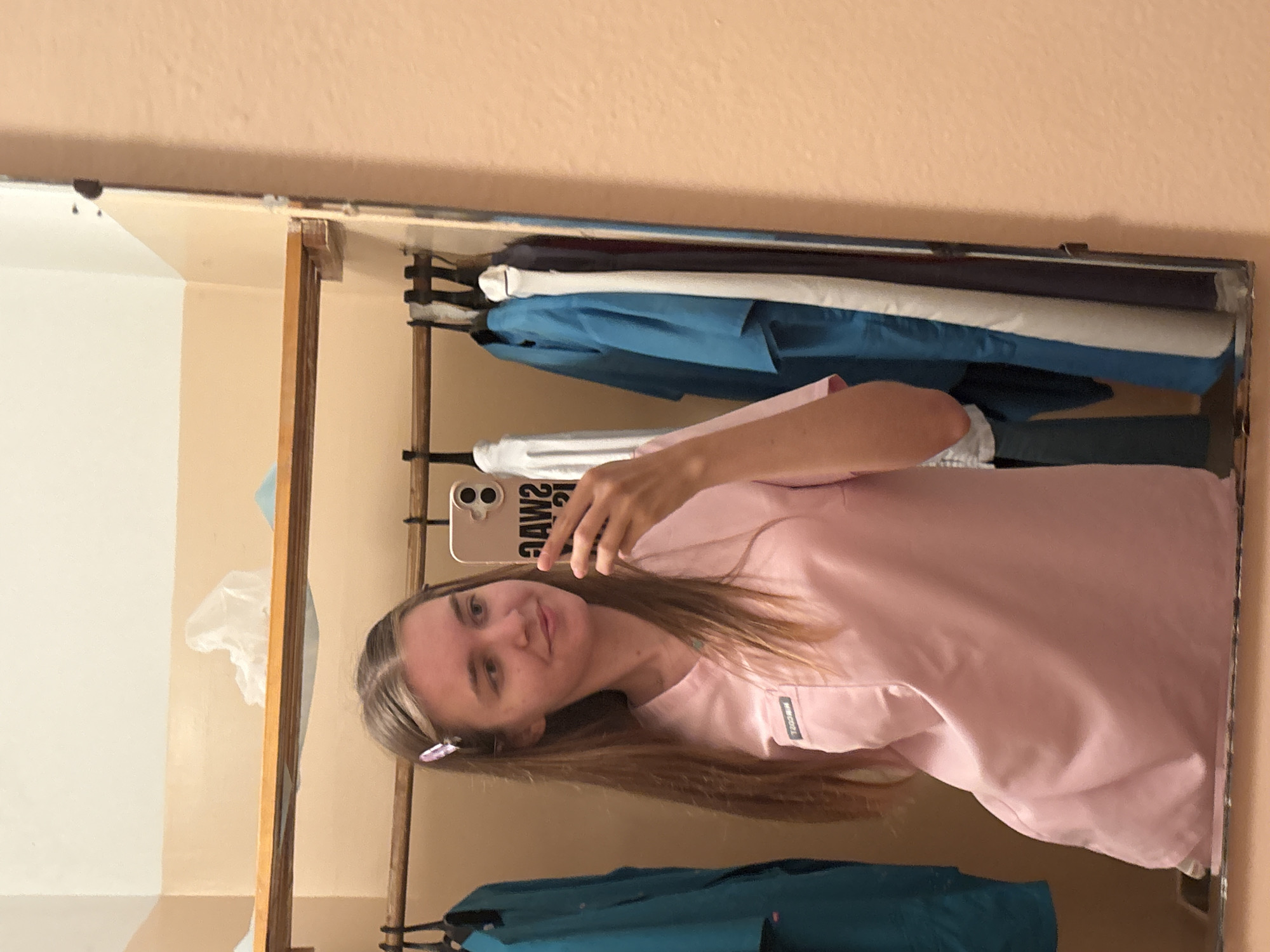 A young Caucasian woman with long, straight brown hair and a pink hair clip is taking a mirror selfie in what appears to be a locker room. She is wearing a pink scrub top. She is holding a smartphone in her right hand, which is angled to capture her reflection. Behind her, on a shelf, are what appear to be folded paper products. A wooden rod is visible, holding several blue and white garments on hangers. The wall to the right of the mirror is a light peach color. The scene is likely in the city of Chaikovsky, Russia, given the provided context.