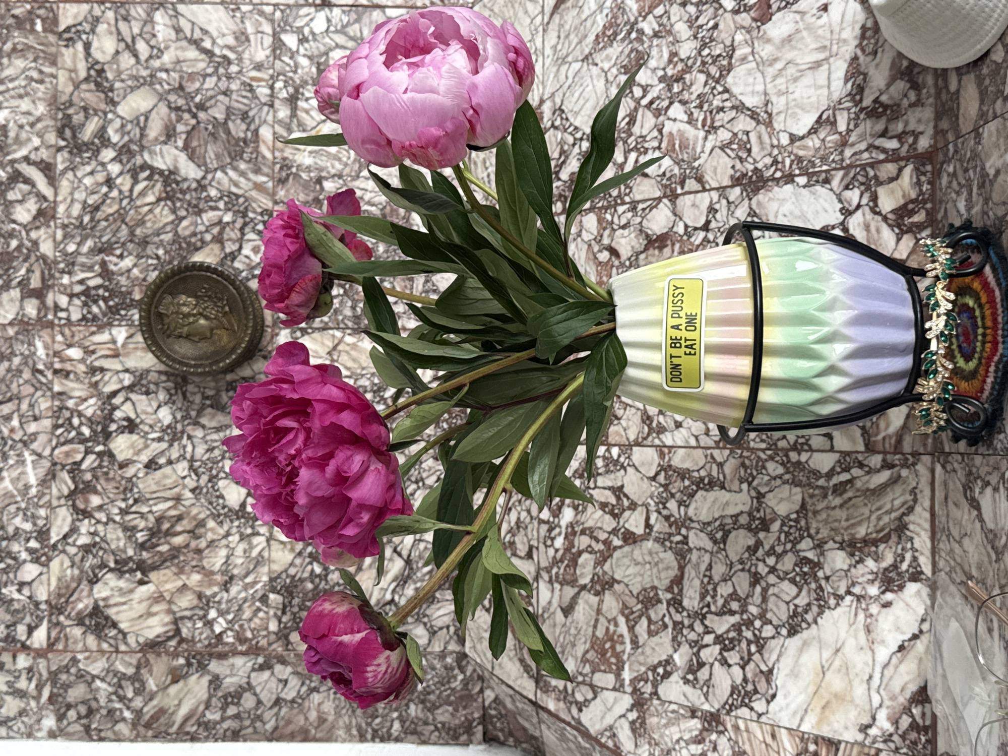The image depicts a still life of a bouquet of pink peonies in a vase, placed on a marbled surface against a marbled wall. A small, circular metal medallion with a profile image is visible on the wall behind the flowers. The vase is a pastel gradient of green, yellow, and purple, with a black metal holder. A yellow sticker on the vase reads "DON'T BE A PUSSY EAT ONE". A decorative element with green jewels and colorful threads is at the base of the vase holder. The scene appears to be indoors, possibly a home or studio. There is no indication of a specific location in Plovdiv, Bulgaria.