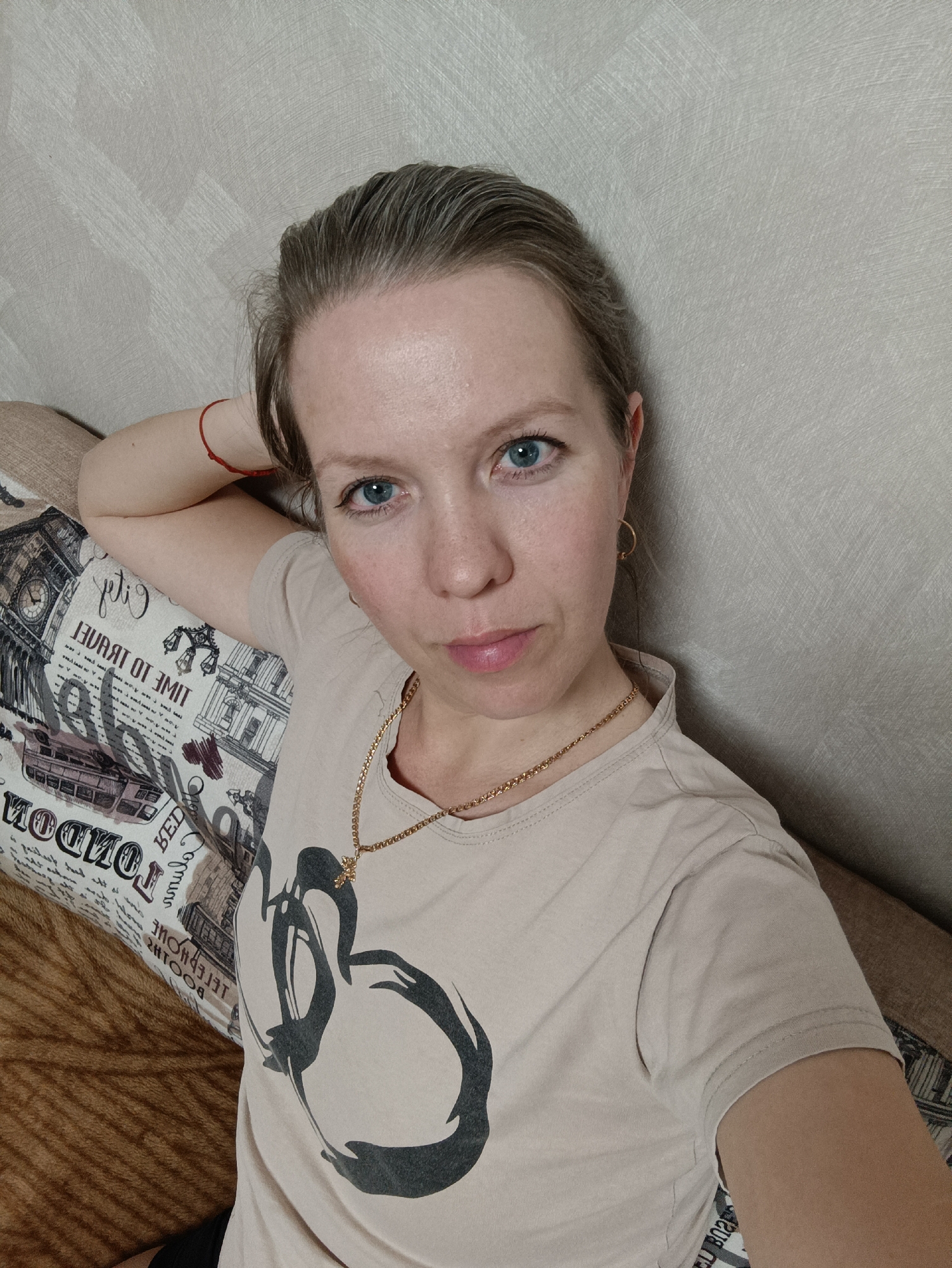 A woman is resting indoors. She has fair skin, blue eyes, and light brown hair pulled back. She is wearing a beige t-shirt with a black swan design, a gold chain necklace, and small gold hoop earrings. A red string bracelet is visible on her left wrist. She is leaning against a decorative pillow with a London-themed print featuring the Big Ben clock tower and text in English. The background is a light beige wall. The environment indicates a domestic setting. The location in the city of Chaikovsky, Russia is not discernible from the image.