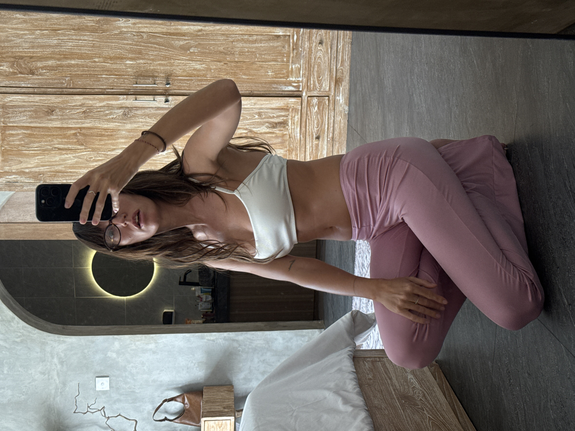A woman in glasses is posing for a selfie in a mirror. She is wearing a white top and pink leggings and is kneeling on the floor. The reflection shows a bedroom with a bed and a bathroom with a round, lit mirror. The reflection shows North Kuta, Indonesia.