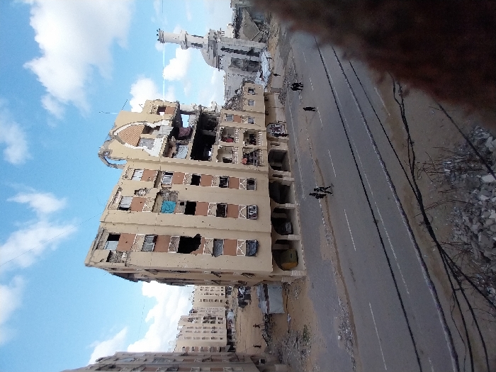 The image depicts a scene of extensive urban destruction in Khan Yunis, Palestinian Territory, under a partly cloudy blue sky. A severely damaged, light brown/beige multi-story residential building dominates the mid-ground, exhibiting large holes in its exterior walls, missing windows, exposed interiors, and significant structural collapse on its upper floors and roof, including a distinctive arched facade. Blue fabric hangs from a fourth-story window. To the right, a heavily damaged mosque with a white minaret and a partially collapsed dome is visible, surrounded by rubble. To the left, several other multi-story residential buildings also show signs of damage, with debris scattered around them. A two-lane asphalt road runs across the mid-ground. On the road, two individuals in dark clothing walk left-to-right, while a single person in dark attire walks right-to-left further down. A small group of two to three individuals stands or walks near the damaged mosque on the far right. Indistinct figures are also visible in the background near the buildings on the left. The ground surrounding the road and buildings consists of dirt and scattered rubble. Dark utility lines and additional rubble occupy the immediate foreground, suggesting an elevated photographic perspective.