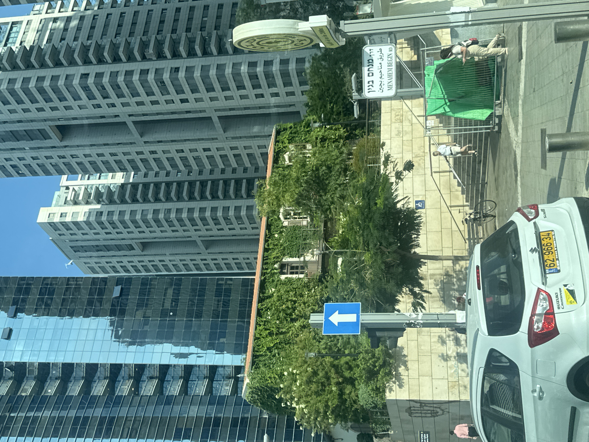 The image shows a street scene in Tel Aviv, Israel, featuring a mix of modern and older architecture, with lush greenery. In the foreground, a white Suzuki crossover is parked, displaying Israeli license plates and stickers. Further back, a building covered in dense green ivy stands out, its red-tiled roof visible. Above and behind this building, several tall, contemporary skyscrapers with glass facades and balconies dominate the skyline. The street includes a sidewalk, a traffic sign with an upward-pointing arrow, stairs leading up to a pathway, and a street sign indicating "MENAHEM BEGIN RD" in both Hebrew and English. People are visible walking on the stairs and sidewalk. A section of green construction fencing is also present near the stairs. The overall environment suggests a sunny day in an urban setting.