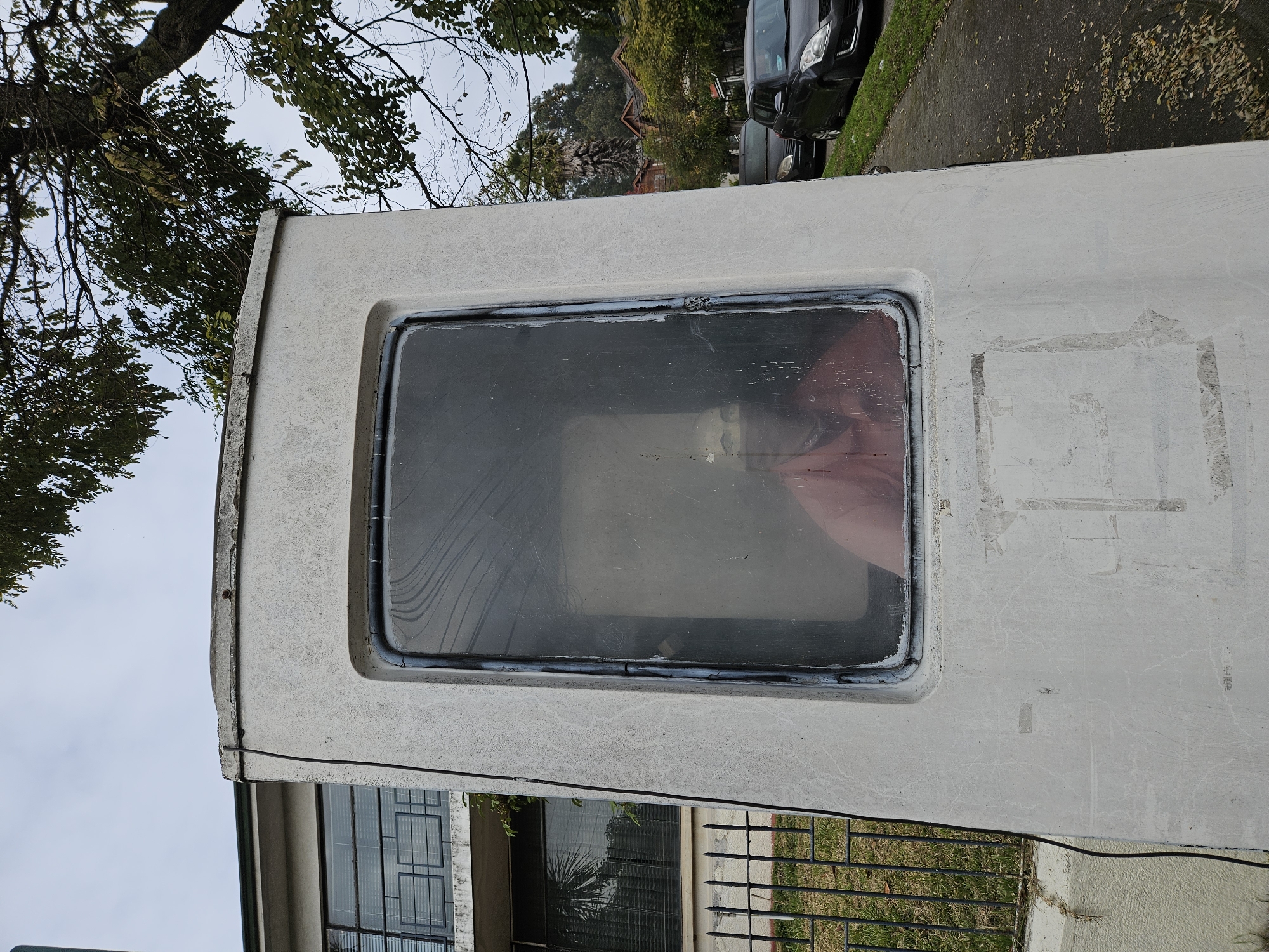 An exterior shot depicts what seems to be the back of a camper van parked on a street. The white exterior of the van is worn, with a prominent window revealing a reflection. The reflection shows a person wearing what appears to be a red garment inside the vehicle. In the background, there's a glimpse of a street with cars parked and some vegetation. Below the van, there is a glimpse of a building with black framed windows and a security fence. Given the context, the location might possibly be in the city of Concepcion, Chile, but definitive confirmation is absent from the image alone.
