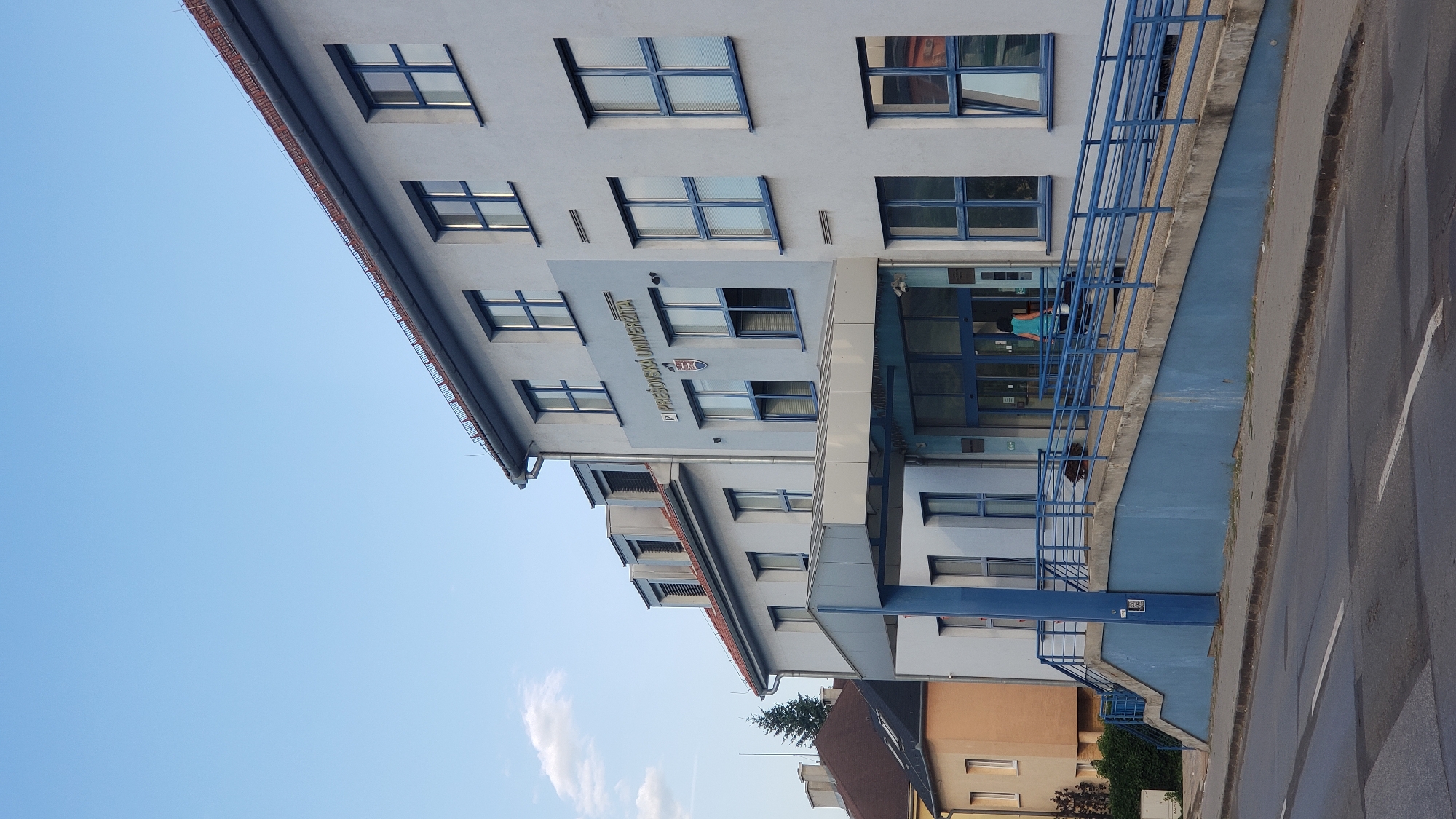 The image depicts the exterior of the University of Prešov (Prešovská univerzita) building. The building is light blue with multiple stories and numerous blue-framed windows. Above the entrance, the university's name and coat of arms are visible. A blue ramp with railings leads up to the entrance. Two individuals, one wearing a turquoise top, are seen on the ramp near the entrance, and a brown dog is visible on the ramp as well. The building is situated on a street with a road and sidewalk in the foreground. The sky is clear blue with a few wispy clouds. To the left of the main building, other structures, including houses with brown roofs and lighter colored walls, are partially visible, suggesting an urban setting within Prešov, Slovakia.