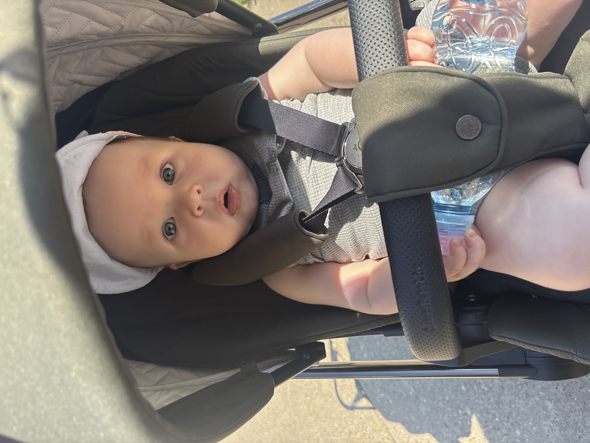 A baby with blue eyes and a white hat is strapped into a dark-colored stroller, holding a water bottle. The stroller is positioned on a sunny surface. The location is specified as Марковский, Russia, but no other details confirm this.