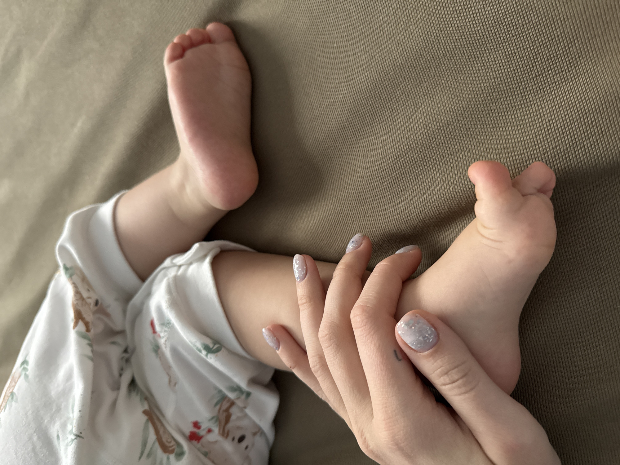 A close-up shot shows a baby's legs and feet on a textured tan surface, possibly a bed or sofa. The baby is wearing white pajamas with a light-colored animal print. An adult's hand with light-colored nails with a glitter design gently touches one of the baby's feet. The photo does not provide information that would determine its location or indicate that it was taken in Moscow, Russia.