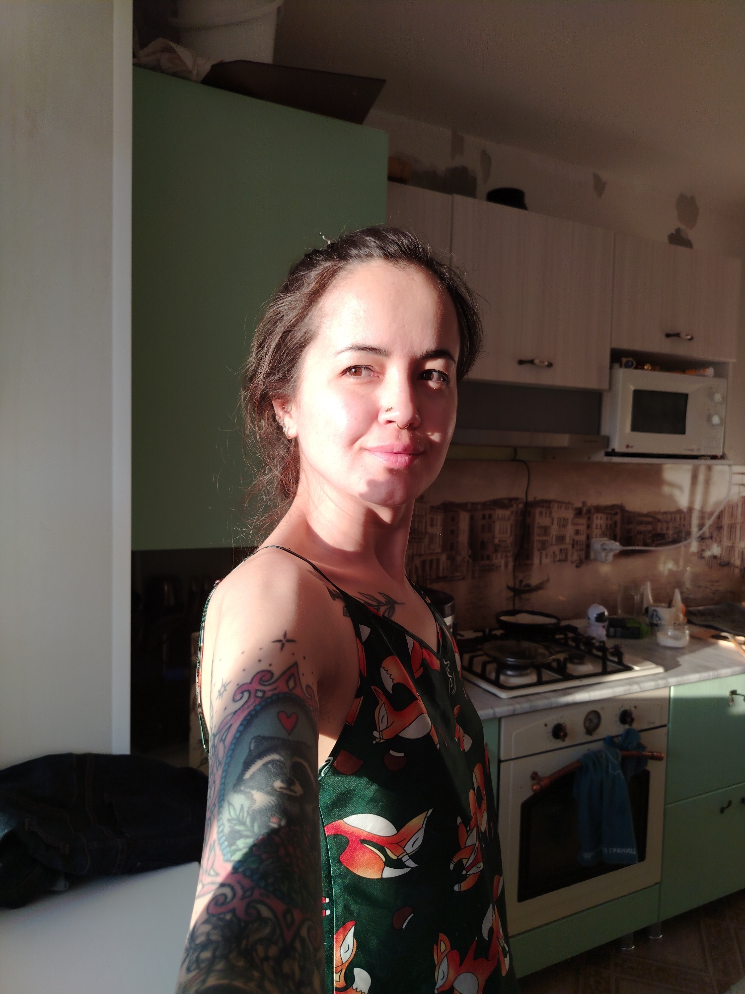 A woman stands in a kitchen. She has dark hair pulled back and is wearing a sleeveless dress with a fox pattern. She has multiple tattoos on her left arm. The kitchen has light green cabinets and a white stove and microwave. The backsplash behind the stove has an image of Venice. There is a dish towel hanging on the oven door. A window to the left of the woman is casting light on her and the kitchen. The location is inferred from the backsplash mural depicting Venice, Italy. The information indicates Noviy, Russia, is related to the subject but does not describe the image directly.