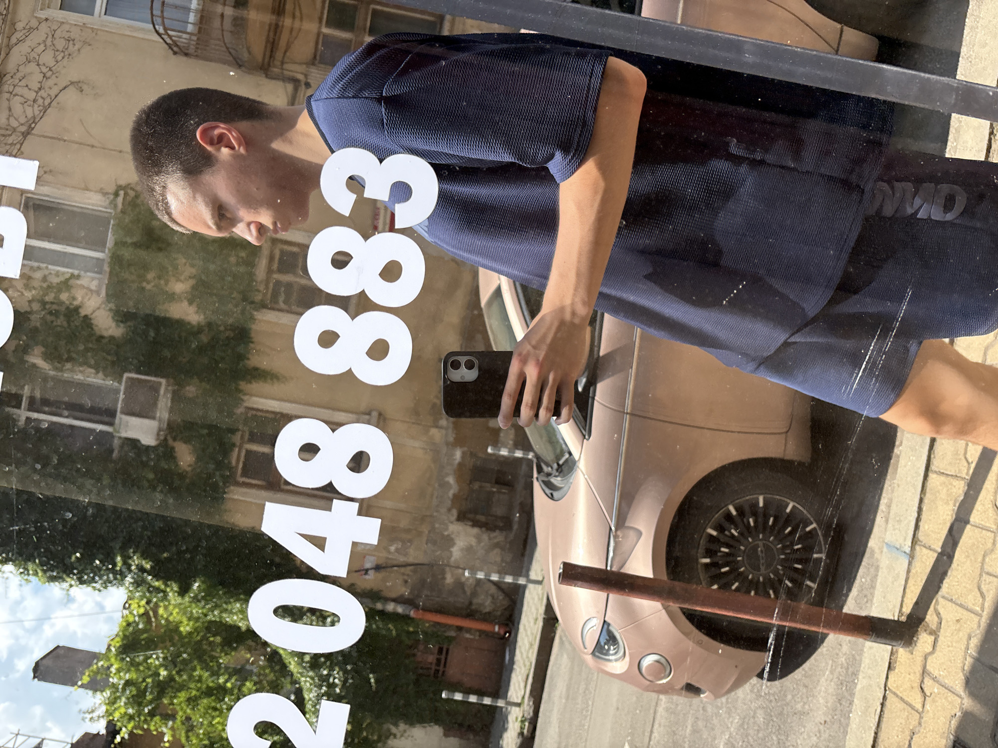 A young male, seen in profile looking right, captures a selfie in a reflective glass surface on a sunny day in Sofia, Bulgaria. He has short dark hair and wears a dark blue, textured short-sleeved shirt and matching shorts with reversed text (appearing as "OMD" or "OMC") visible on the left leg. He holds a black smartphone, likely an iPhone with two rear cameras, in his right hand.
The reflection reveals a street scene. A light brown/rose gold colored Fiat 500 is parked on the street, with its front left section and a black multi-spoke wheel with white accents visible. The street features tan interlocking pavement stones and several white and black striped bollards. A reddish-brown pole stands in the immediate foreground, partially obscuring the car's wheel and the person's lower body.
In the background, old multi-story buildings line the street. One building facade is partially covered in dense green ivy, and an air conditioner unit is mounted on its wall. Other details include various windows, a balcony railing on an adjacent building, and a brown door/gate further down the street. Overhead, a blue sky with scattered white clouds is visible.
Adhered to the reflective glass surface are large white vinyl numbers: "2048 883". Above these numbers, partial white letters, possibly "Y" or "BY", are also visible, inverted due to the camera angle and reflection. The number format is consistent with a Bulgarian telephone number, with "2" potentially indicating the Sofia area code.