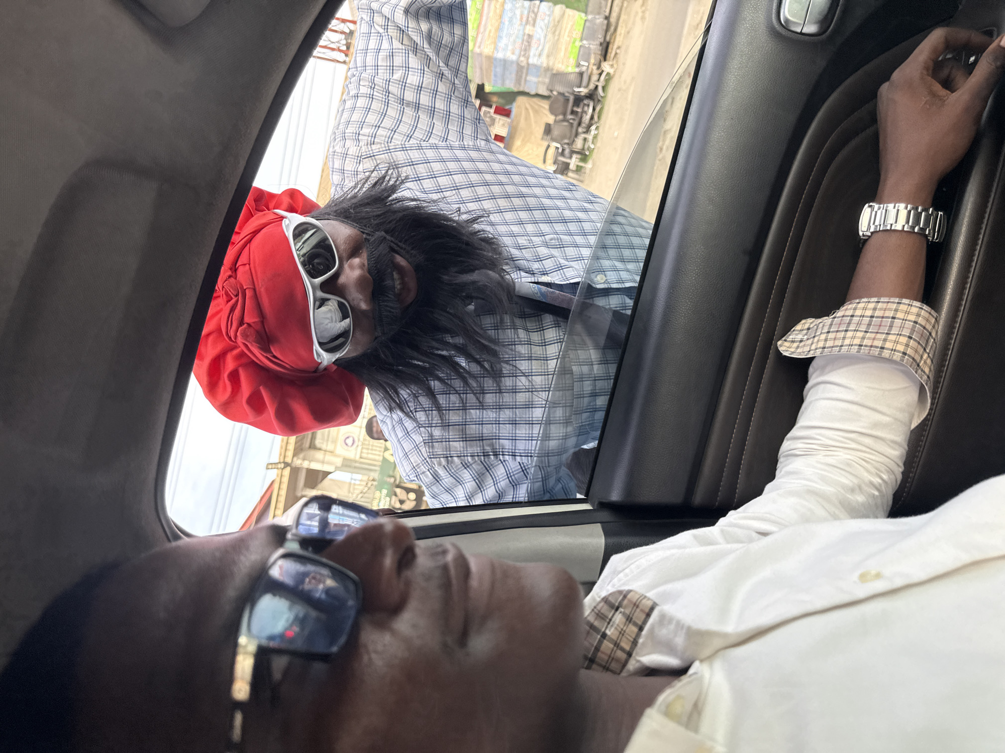 From the perspective of a person inside a vehicle, the image shows another man outside the car window. The man outside is wearing a red head covering, white sunglasses, and a blue and white plaid shirt. He has long black hair. The person inside the vehicle is wearing glasses, a white long-sleeved shirt with a plaid cuff, and a silver watch. The interior of the vehicle has dark upholstery. Based on the appearance of the surroundings, it is likely in Lagos, Nigeria.