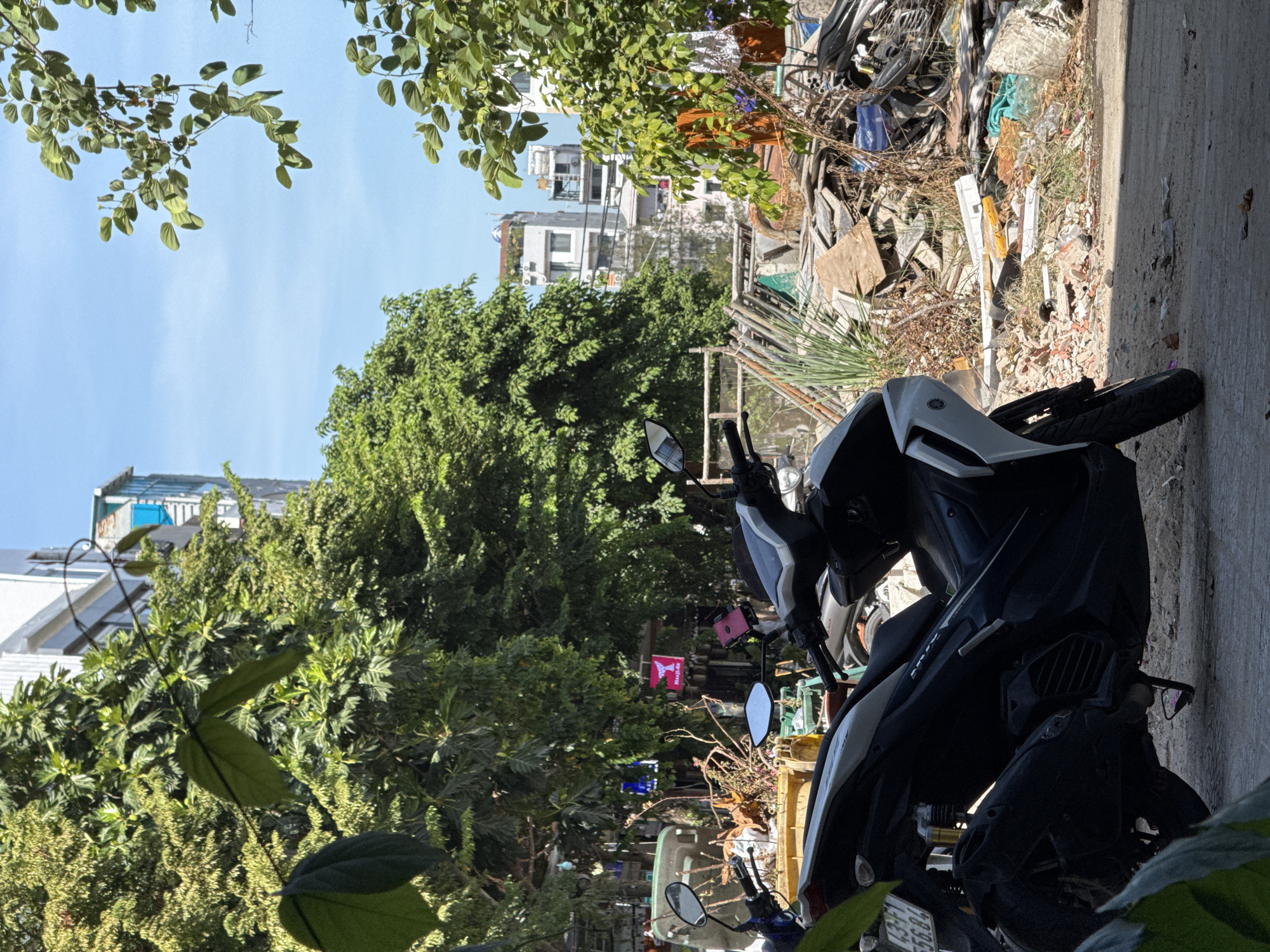 In Đà Nẵng, Vietnam, an outdoor scene unfolds with a focus on a parked black and white scooter in the foreground. The scooter, a Yamaha NVX 155, is positioned on a dirt and paved surface, partially obscured by lush green foliage. Behind the scooter, a chaotic jumble of discarded building materials, including broken tiles, wood scraps, and various debris, dominates the middle ground. In the distance, several multi-story buildings stand against a clear blue sky, suggesting an urban environment. A large, dense green tree is prominent to the left of the frame, contributing to the natural elements within the cityscape. A few signs are visible among the debris, one pink with a whale tail logo. The overall impression is one of a cluttered, lived-in urban space with a blend of natural and man-made elements.