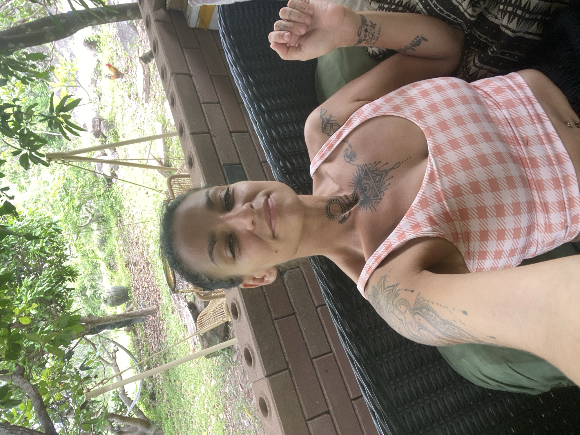 A woman with dark hair pulled back is taking a selfie while sitting on a wicker chair. She is wearing a pink and white checkered tank top and has multiple tattoos on her chest, neck, and arms. The background shows a lush, green, outdoor setting with trees, foliage, and what appears to be a brick wall and part of a building. There are also some rustic-looking structures in the background, possibly a hammock and a swing. The overall impression is of a relaxed, natural environment. The location is identified as Mũi Né, Vietnam.