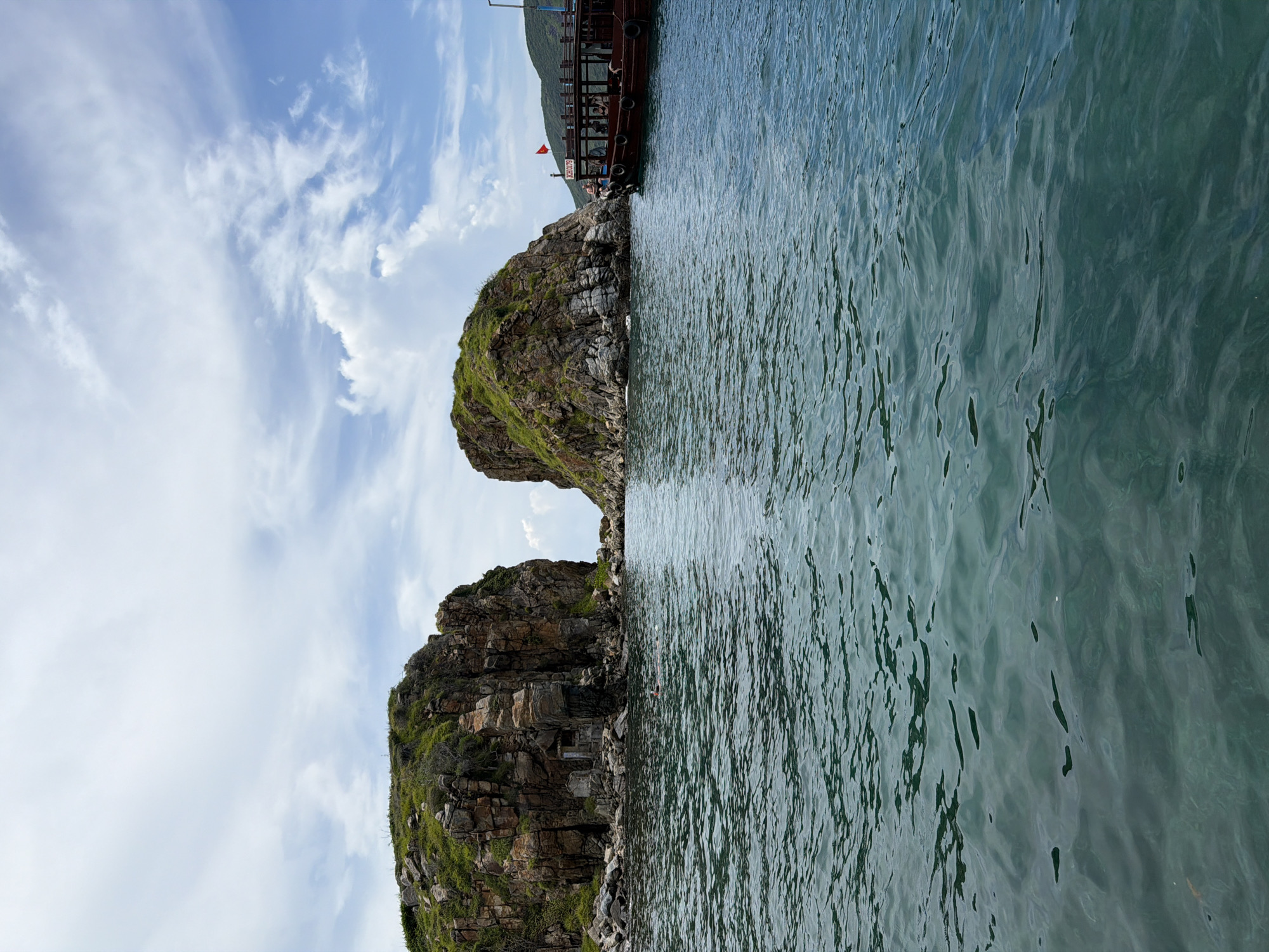 The image shows a view of the sea with two rocky islands. The islands are covered with green vegetation and are located close together, creating a narrow passage of water between them. The water is rippled and reflects the sky. A boat with people on board is docked at the edge of one of the islands. The sky is blue with scattered white clouds. The image was taken in Bac Nha Trang Ward, Vietnam.