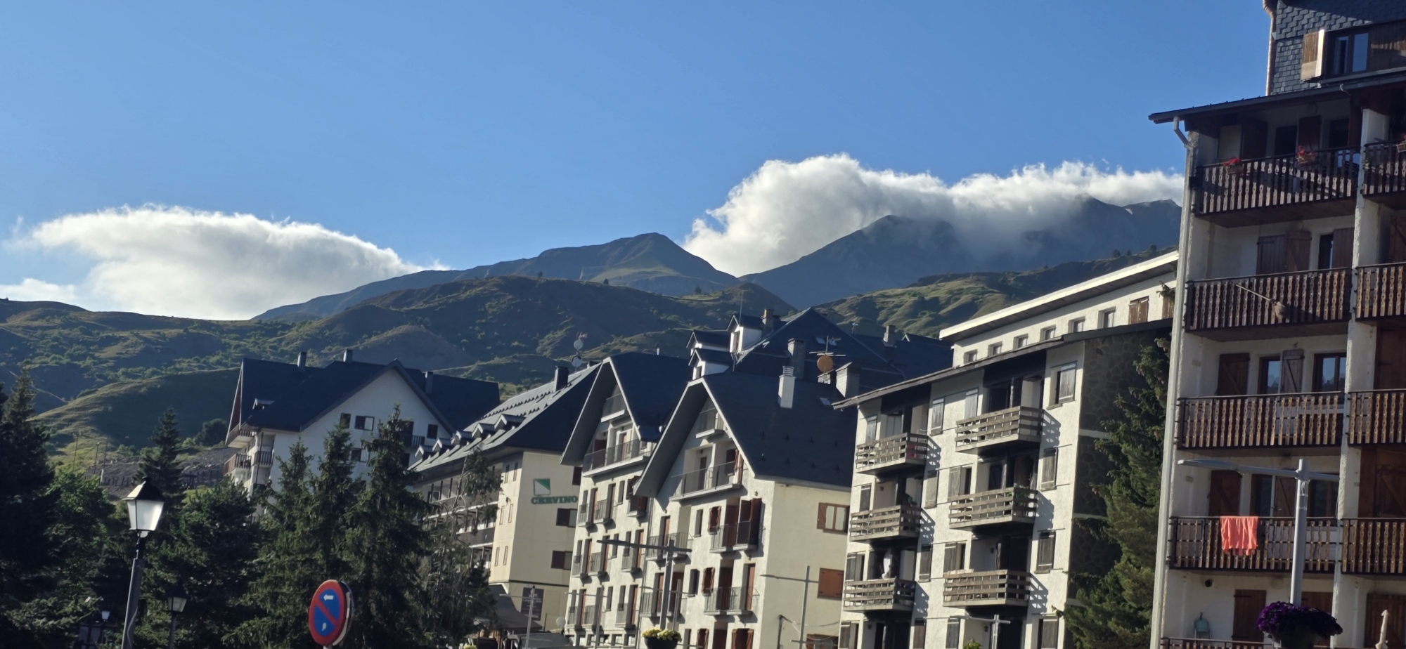 The image depicts a view of apartment buildings in Sallent de Gállego, Spain, with a mountainous landscape in the background. The buildings are multi-story structures with dark-tiled roofs and balconies. Some of the balconies have wooden railings and shutters. The architecture suggests a mountain resort town. The foreground features evergreen trees and a lamppost. A circular red and blue sign is visible on the left side of the image. The sky is a clear blue with some white clouds hovering over the mountain peaks. The overall scene is bright and suggests a pleasant, sunny day.