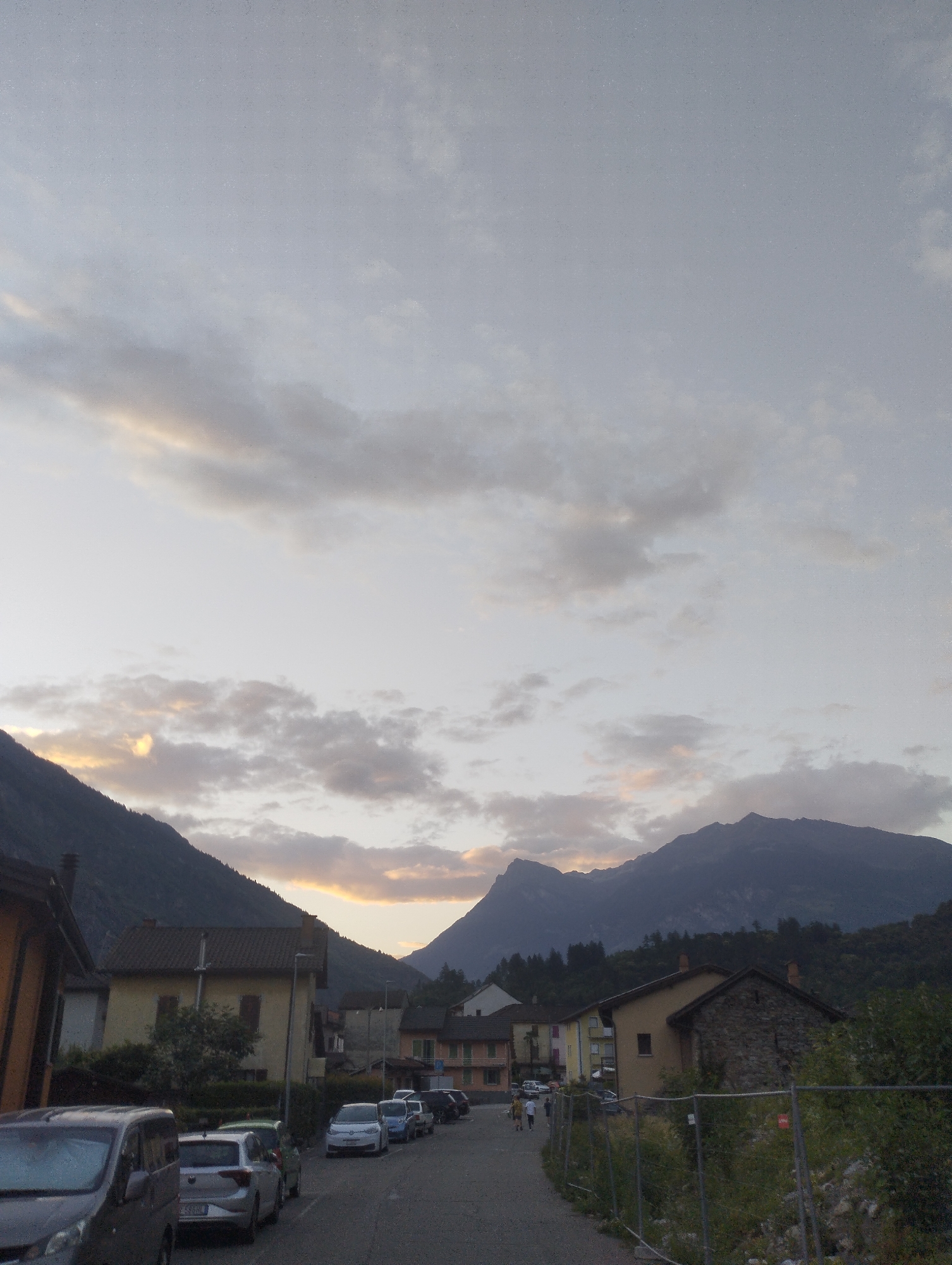 The image depicts a street in a village with mountains in the background. The sky is overcast with scattered clouds, tinged with the warm hues of either sunrise or sunset. Several cars are parked along the side of the road. On the left, a dark-colored van is parked, followed by a silver hatchback. Further down the street, other vehicles are visible, including a white hatchback and a blue sedan. Buildings line the street, with a mix of architectural styles. Some structures appear to be residential homes, while others may be commercial or public buildings. The overall scene suggests a quiet, possibly rural, setting at either the beginning or end of the day.