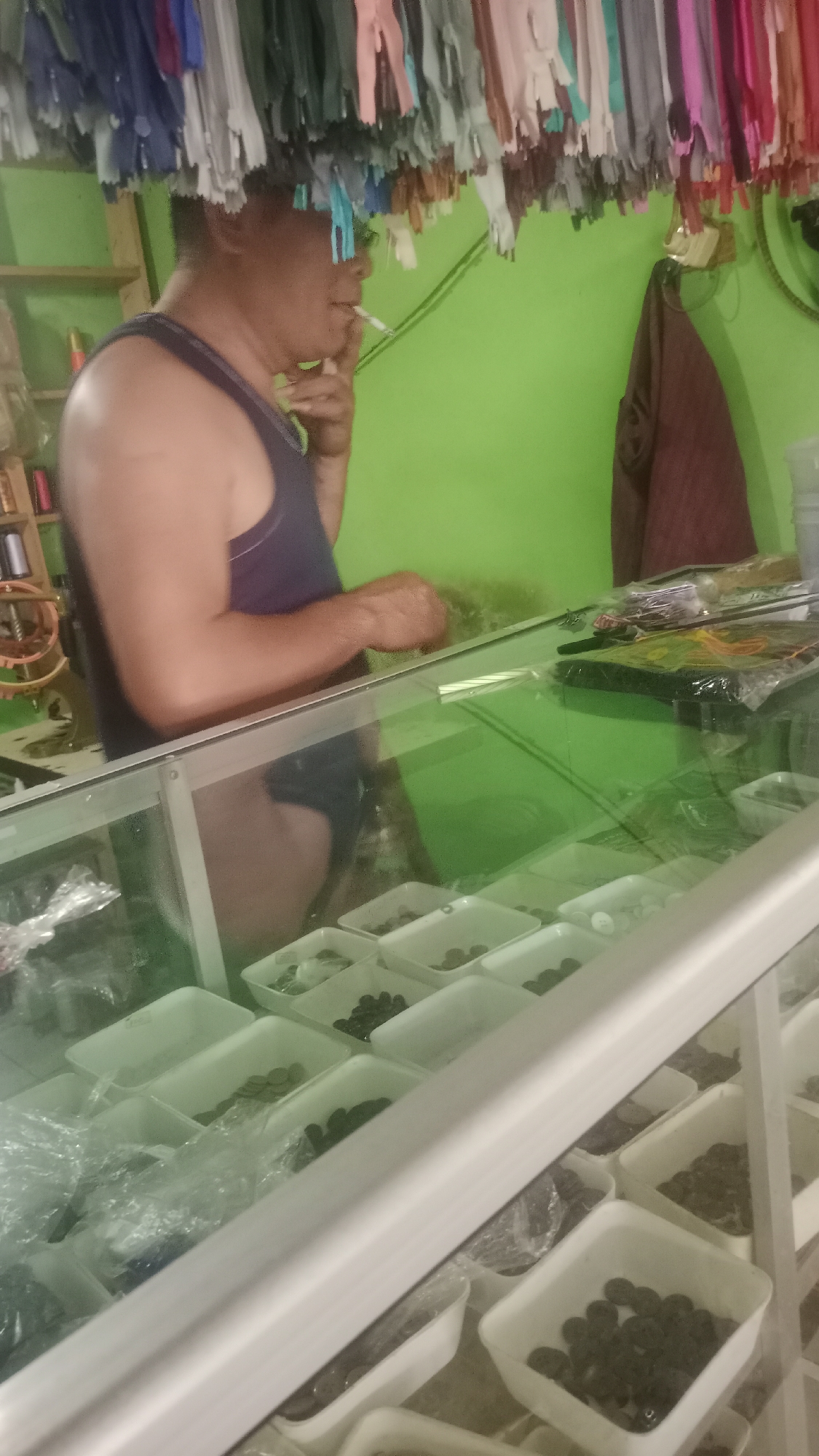 In a dimly lit shop in Cogreg, Indonesia, a person wearing a dark tank top and underwear stands behind a glass counter, smoking a cigarette. Above them, an assortment of colorful zippers hangs from the ceiling. The counter is filled with various small items in white plastic containers, some of which appear to be beads or buttons. The shop has a vibrant green wall and shelves with spools of thread in the background. A brown garment hangs on the wall to the right of the person. The overall impression is one of a small, cluttered shop.