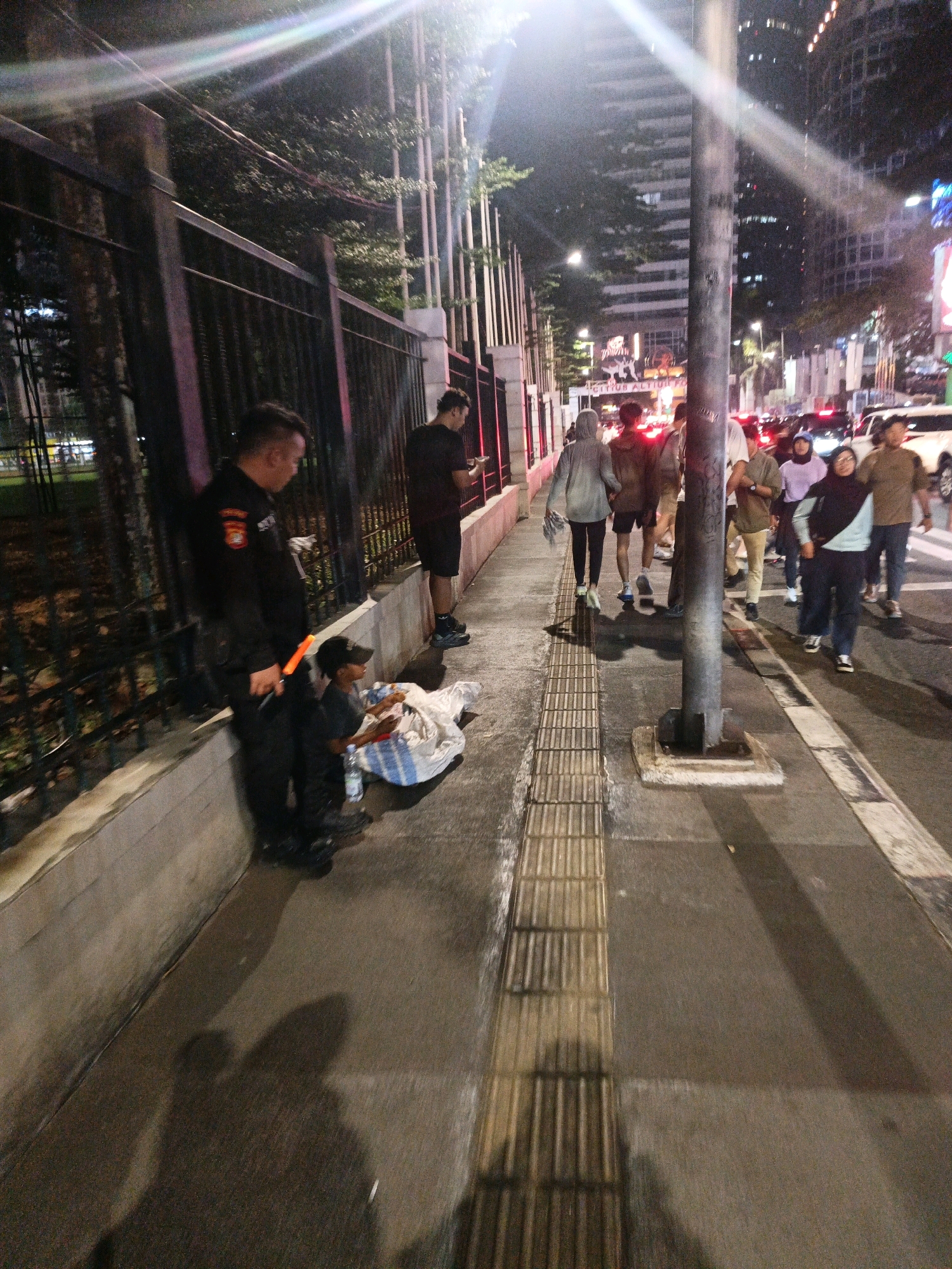 A security guard in uniform stands next to a sidewalk in Central Jakarta, Indonesia, by a black metal fence. He is looking down at a young person sitting on the ground next to a pile of white and blue bags. The young person is wearing a black cap and appears to be sorting through the bags. Other pedestrians are walking by on the sidewalk, with a busy street and city buildings in the background. The scene is illuminated by streetlights and the lights from passing vehicles.