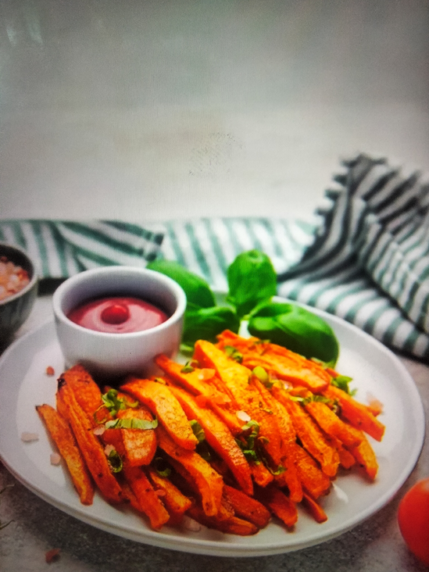 A plate of sweet potato fries is served with a side of ketchup and garnished with chopped green onions. The plate is white and sits on a grey surface. In the background, there is a green and white striped cloth, and some basil leaves. There is no indication of the city of Payagaboh, Indonesia in this image.
