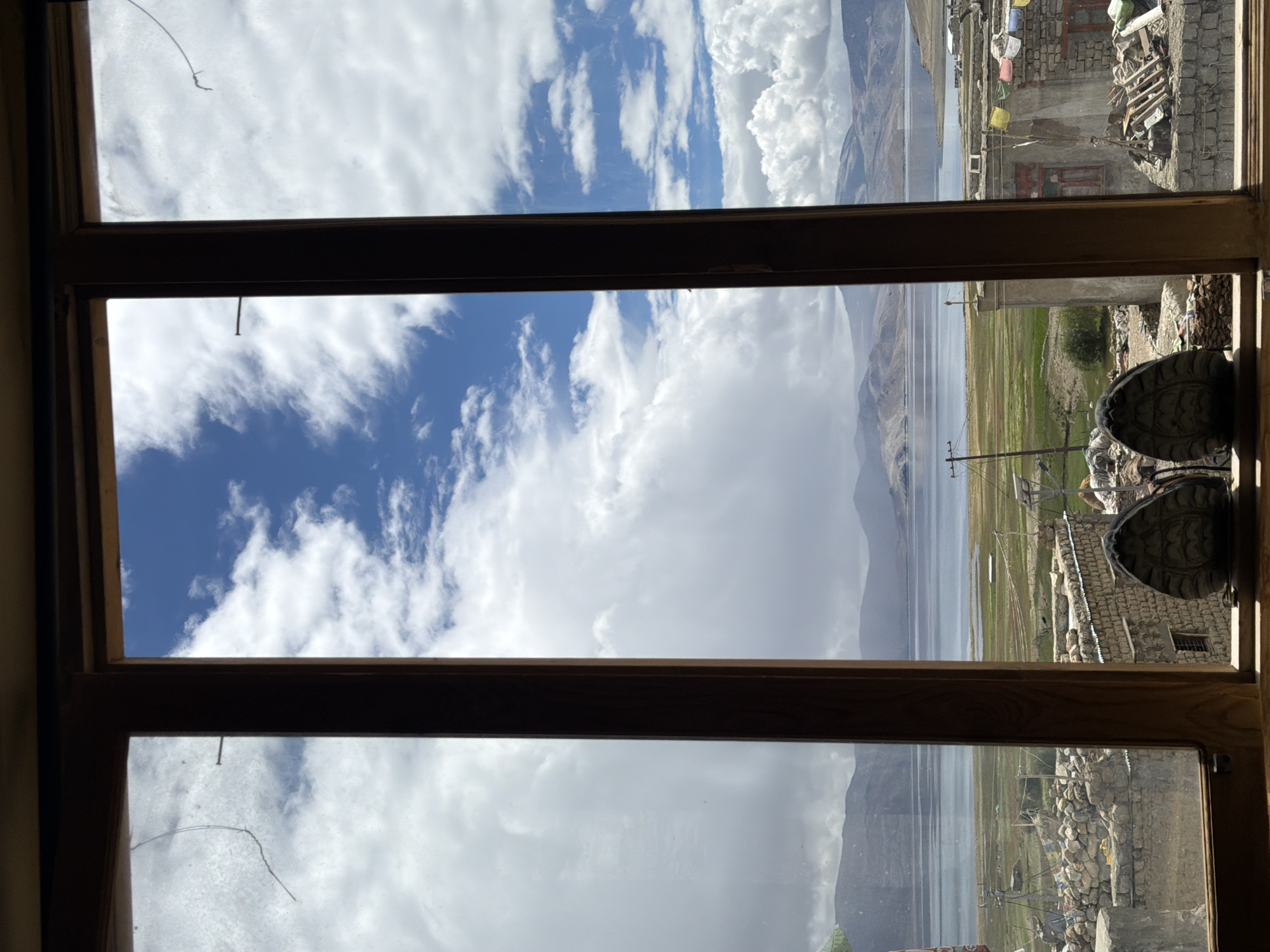 This image captures a view through a wooden window frame at a scene in Karzok, India. The window is divided into three vertical panes. The foreground of the windowsill shows two hiking boots positioned side-by-side. Beyond the window, a wide expanse of a lake stretches towards distant, undulating mountains. The water is a calm, deep blue, reflecting the sky. The landscape surrounding the lake is a mix of green and brown terrain, with some scattered vegetation and a few structures. In the middle ground, there are low-lying buildings constructed from stone and cement, some appearing to be residences with colorful prayer flags strung across them. A few utility poles with wires are also visible. The sky above is a dramatic mix of bright blue and large, fluffy white clouds, suggesting a partly cloudy day. The overall scene depicts a natural landscape with a touch of human settlement in a remote, high-altitude region.