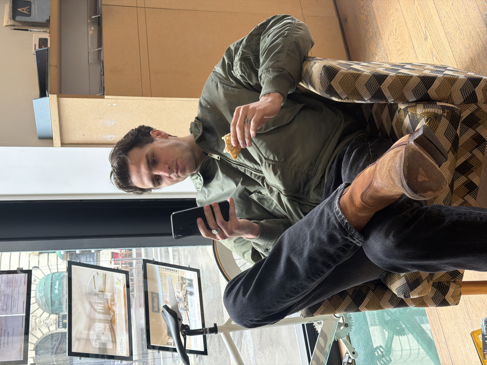 A man sits in a patterned chair, eating something and looking at a smartphone. He is wearing a green jacket, black jeans, and brown boots. A white bicycle is nearby. Through the window, advertisements are displayed, and the architectural details of a Parisian building are visible. This suggests the scene is likely inside a shop or cafe in Paris, France.