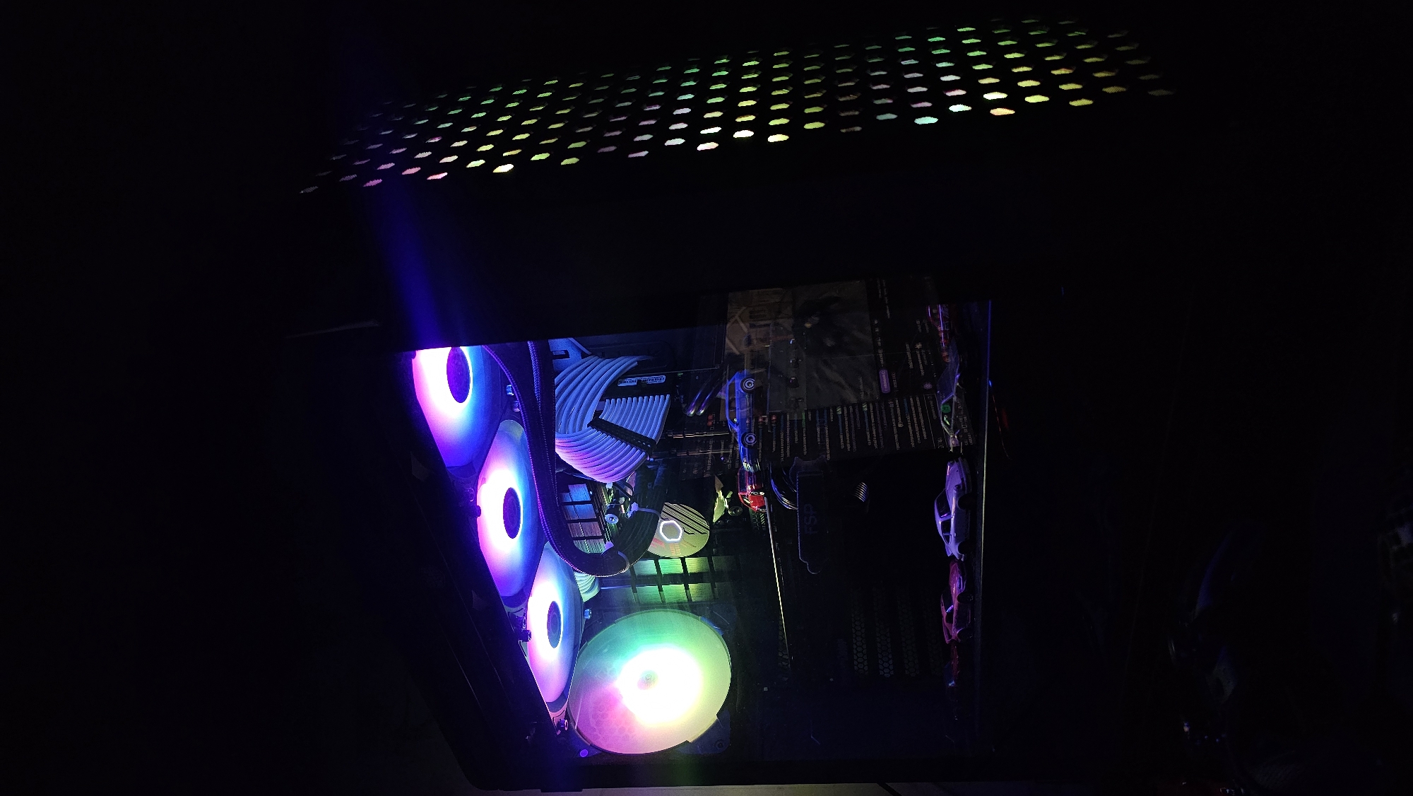 The image depicts the interior of a personal computer case with LED-lit components. Visible components include multiple cooling fans with colorful RGB lighting, white power cables, a motherboard, and possibly a graphics card. Small toy cars are visible in the lower right of the computer case. A section of the perforated top of the computer case is visible in the upper portion of the image. The scene is poorly lit, with the RGB lights providing the main source of illumination. The location is not identifiable from the image, nor can it be placed in Varna, Bulgaria.