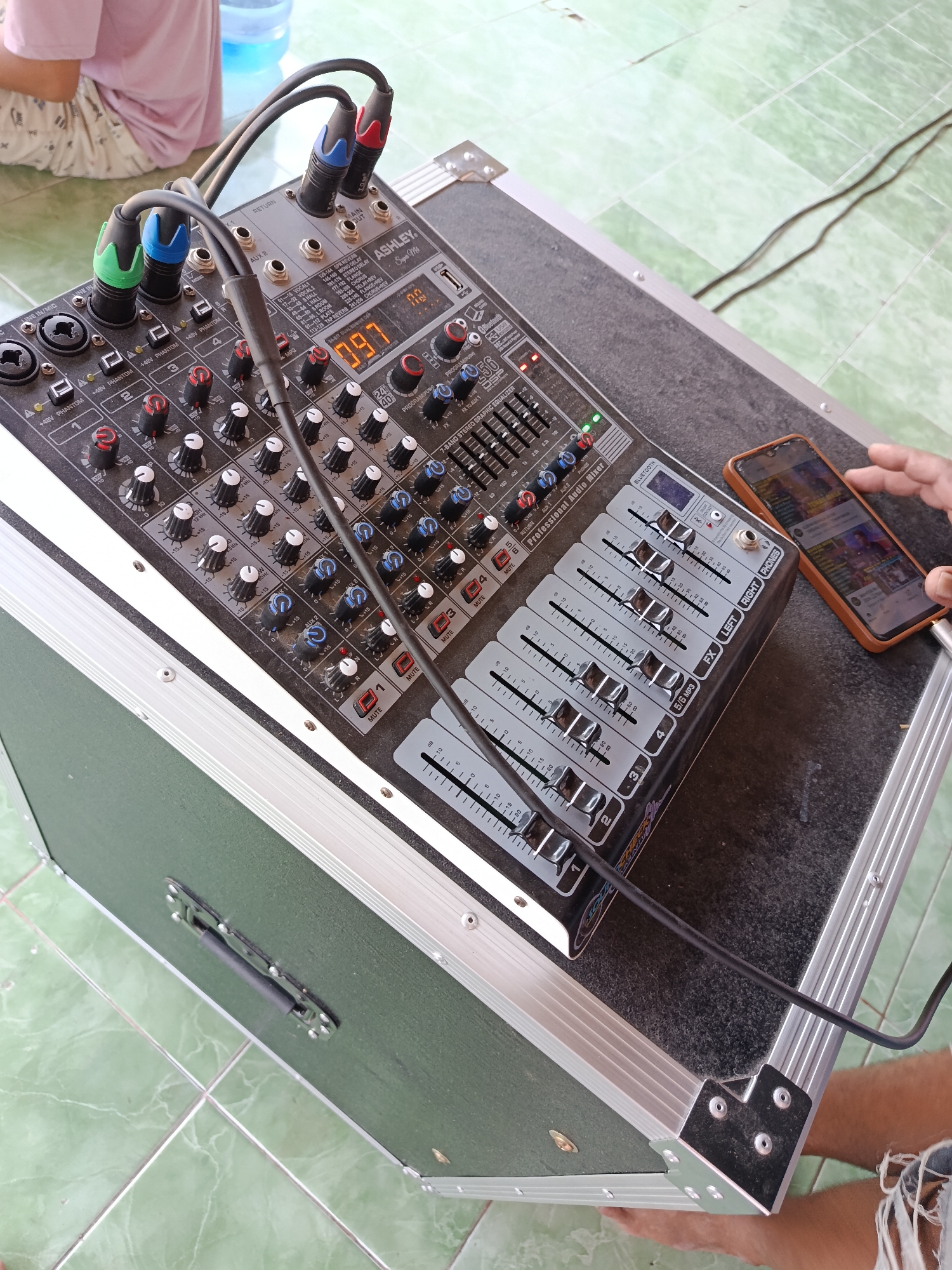 In Jurug, Indonesia, an audio mixer is set up on a dark, textured case. Cables connect to the mixer, which has several knobs and sliders. A smartphone, resting on the case, is being operated by a person's hand. In the background, a person wearing pink and white clothing is visible. The scene is set against a backdrop of light green tiled flooring.