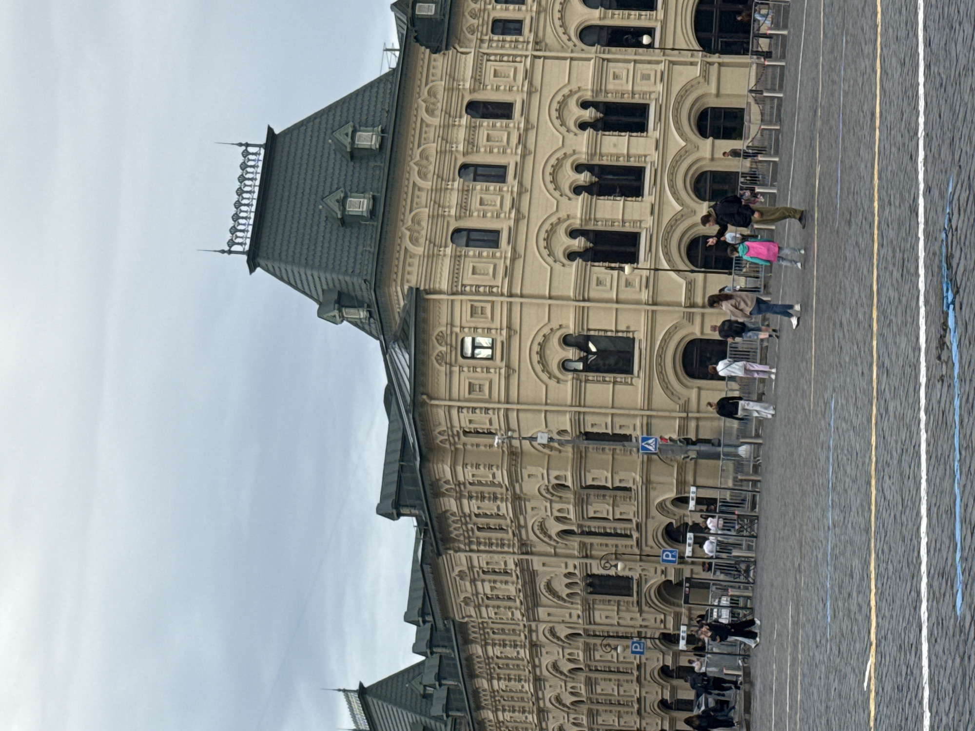 The image shows the facade of a grand building, likely in Moscow, Russia, with an ornate architectural style. The building has multiple stories with numerous windows, many of which are arched. The facade is intricately decorated with detailed carvings and patterns. A dark, steeply pitched roof with dormer windows caps the building.
In the foreground, a cobblestone ground stretches out, marked with colored lines that suggest a designated path or area, possibly for an event. Several people are visible walking along this path, some in small groups and others individually. They appear to be casually strolling or moving through the area. One person in the mid-ground is wearing a bright pink vest, while others are dressed in casual clothing.
Fencing and barriers are set up along the side of the building, indicating a controlled access area or a viewing point for an event. There are also some lampposts and signage, including blue signs with a white "P" symbol, likely indicating parking or a designated zone. The sky is overcast, suggesting a cloudy or possibly early morning/late afternoon time of day. The overall scene appears to be a public space with people going about their day near a significant historical or governmental building. There are no overt signs of a specific event like a parade or festival, but the setup with fencing implies some form of organized activity may have occurred or is expected.