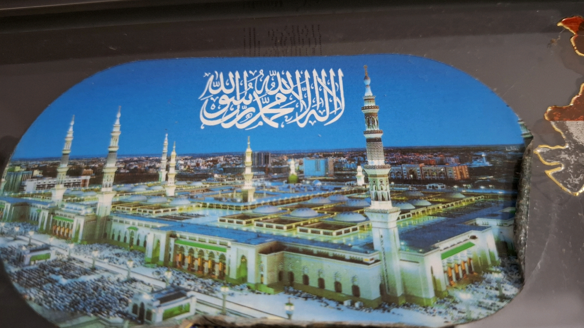 The image depicts a panoramic view of a mosque with multiple minarets and domes, likely representing an important Islamic architectural site. The mosque is surrounded by buildings and appears to be a bustling area with many people gathered, suggesting a place of worship or a significant cultural landmark. Arabic calligraphy is prominently displayed at the top of the image, above the mosque structure. The scene is set against a bright blue sky. The image is framed by a dark, textured border. While the image shows a mosque and people, there is no indication that this location is in Manikkoth, India.