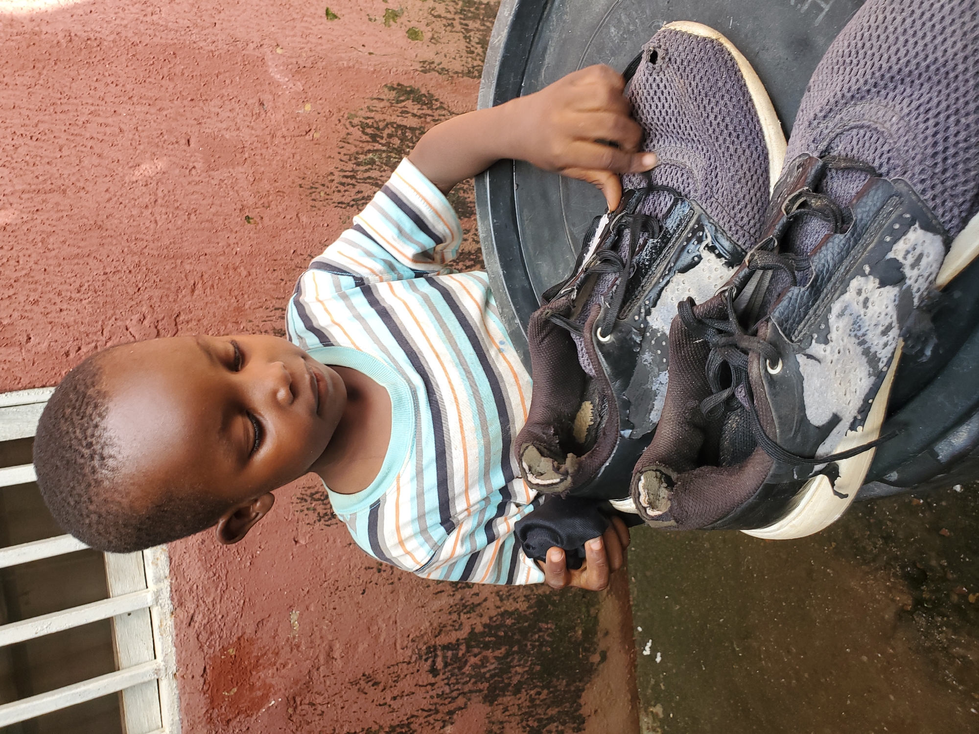 A young boy with dark skin and short black hair is focused on cleaning a pair of well-worn, dark gray athletic shoes. He is wearing a striped t-shirt with horizontal bands of white, blue, orange, and black. The boy is seated outdoors in front of a textured, reddish-brown wall, with a white barred window visible in the upper left corner. The ground appears wet and muddy, with some debris. The shoes are resting in a large, black plastic container. The boy is holding a black cloth in his left hand, and his right hand is touching one of the shoes. The shoes themselves show significant signs of wear, with torn material and visible foam padding. The overall scene suggests a domestic, outdoor setting, likely a courtyard or back alley in Jos, Nigeria.