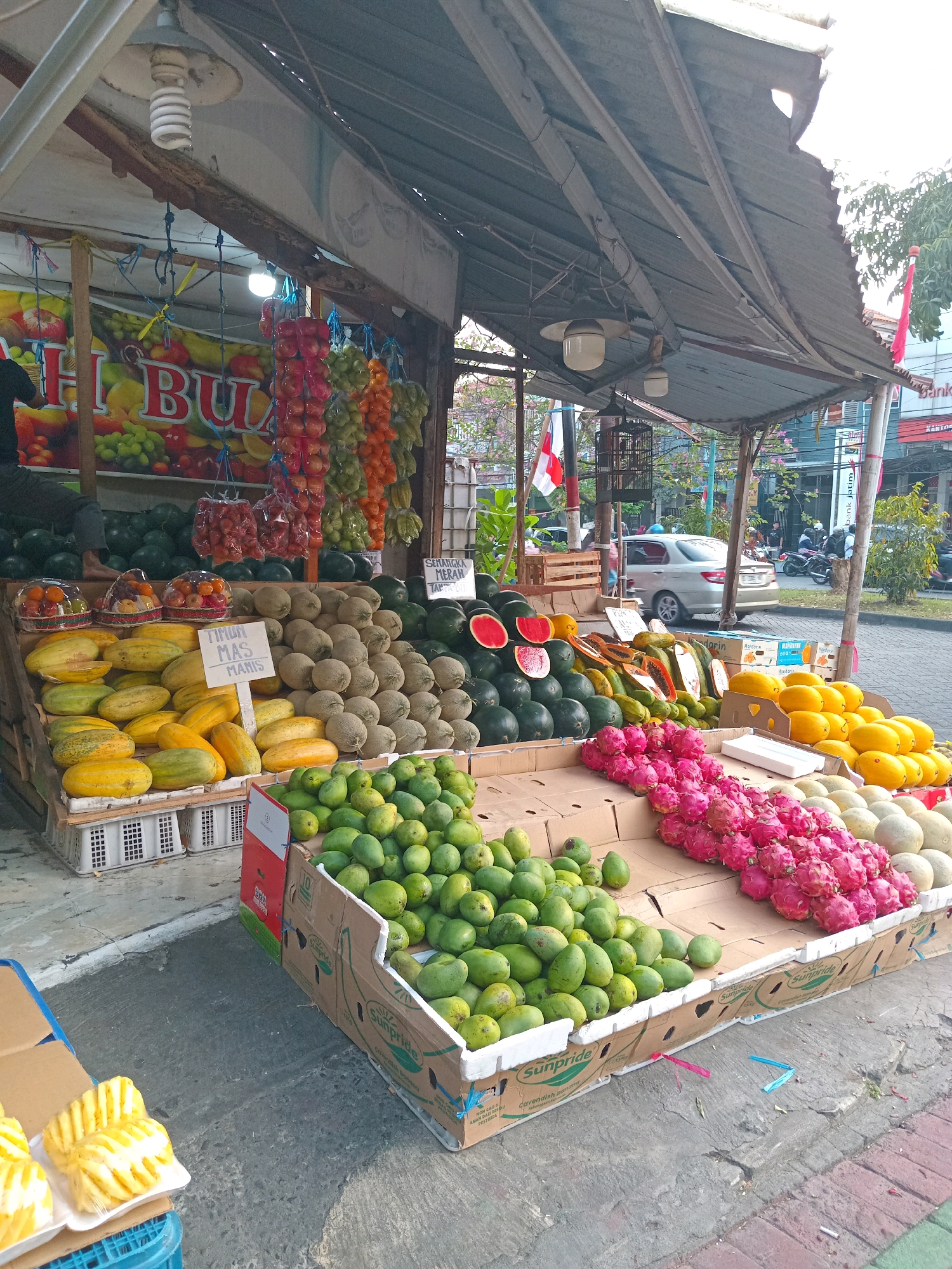 An outdoor fruit stand is displayed in Randuagung, Indonesia. The stand features a variety of fruits, including cantaloupes, watermelons, mangoes, dragon fruit, pineapples, and various types of melons. Some fruits are whole, while others are sliced. The stand is made of wood and is covered by a corrugated metal awning. In the background, a road with passing vehicles and buildings can be seen. There are two Indonesian flags displayed on poles, one partially obscured by the awning. A man is sitting at the stand, possibly the vendor, with his legs crossed. There are also some pre-packaged fruit items on display in blue crates. The ground is paved with concrete.