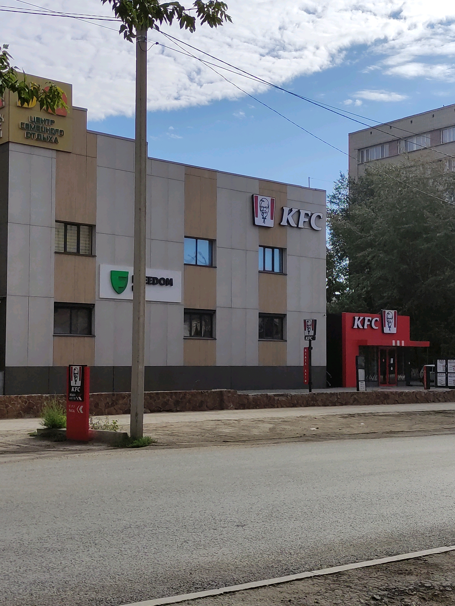 A street-level, eye-level, medium shot captures a section of a modern, multi-story building with a KFC restaurant prominently featured on the right side of the frame. The building has a facade of light-colored panels with vertical stripes of a tan color. Above the KFC entrance, a large "KFC" logo with the Colonel Sanders image is visible. To the left of the KFC, a sign with "KFC" and arrows, likely for a drive-thru, stands on a red pole. Further to the left, a beige building section bears the Cyrillic text "ЦЕНТР СЕМЕЙНОГО ОТДЫХА" (Center for Family Recreation) in dark letters. Below this, a white sign with a green shield logo and the word "FREEDOM" is displayed. Several windows are visible on the building.
The environment includes a paved street in the foreground with a strip of sidewalk or verge along the building. A concrete utility pole stands in the middle of the frame, with overhead wires stretching across the blue sky with scattered white clouds. The building to the far right is a darker, blockier structure with multiple windows. Trees are visible behind the buildings. The scene appears to be a commercial area in the city of Semey, Kazakhstan. No people are clearly visible or identifiable in the frame. The lighting suggests it is daytime, likely under a partly cloudy sky.