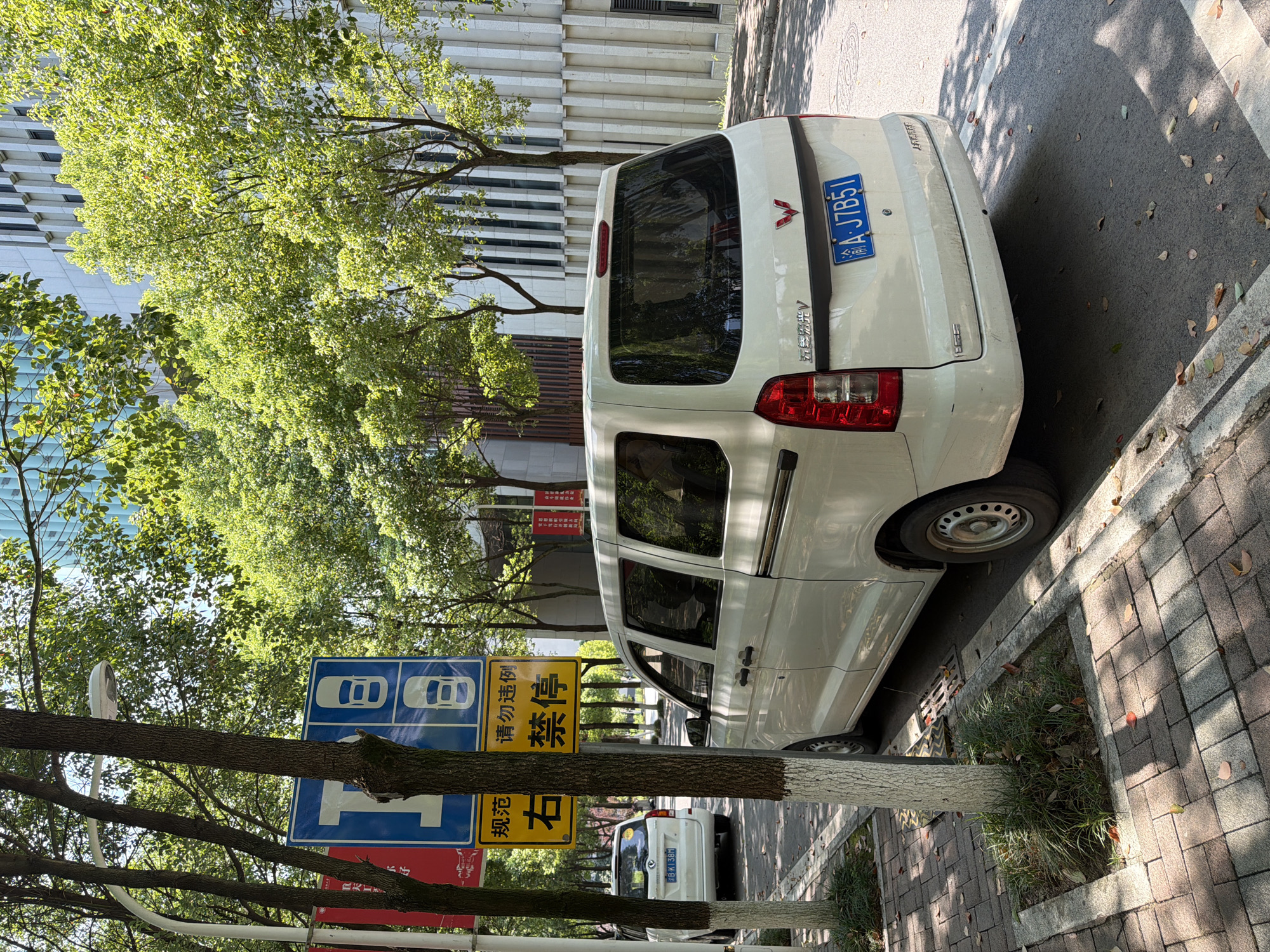 The image shows a street in Jiangjin, China, with two white minivans parked along the side. The street is paved with brick, and there are trees and a modern building in the background. Traffic signs are visible, including one with icons of cars and another with Chinese characters. The license plate of the minivan in the foreground reads "A·J7B51". The overall scene depicts a typical urban street environment on a sunny day.