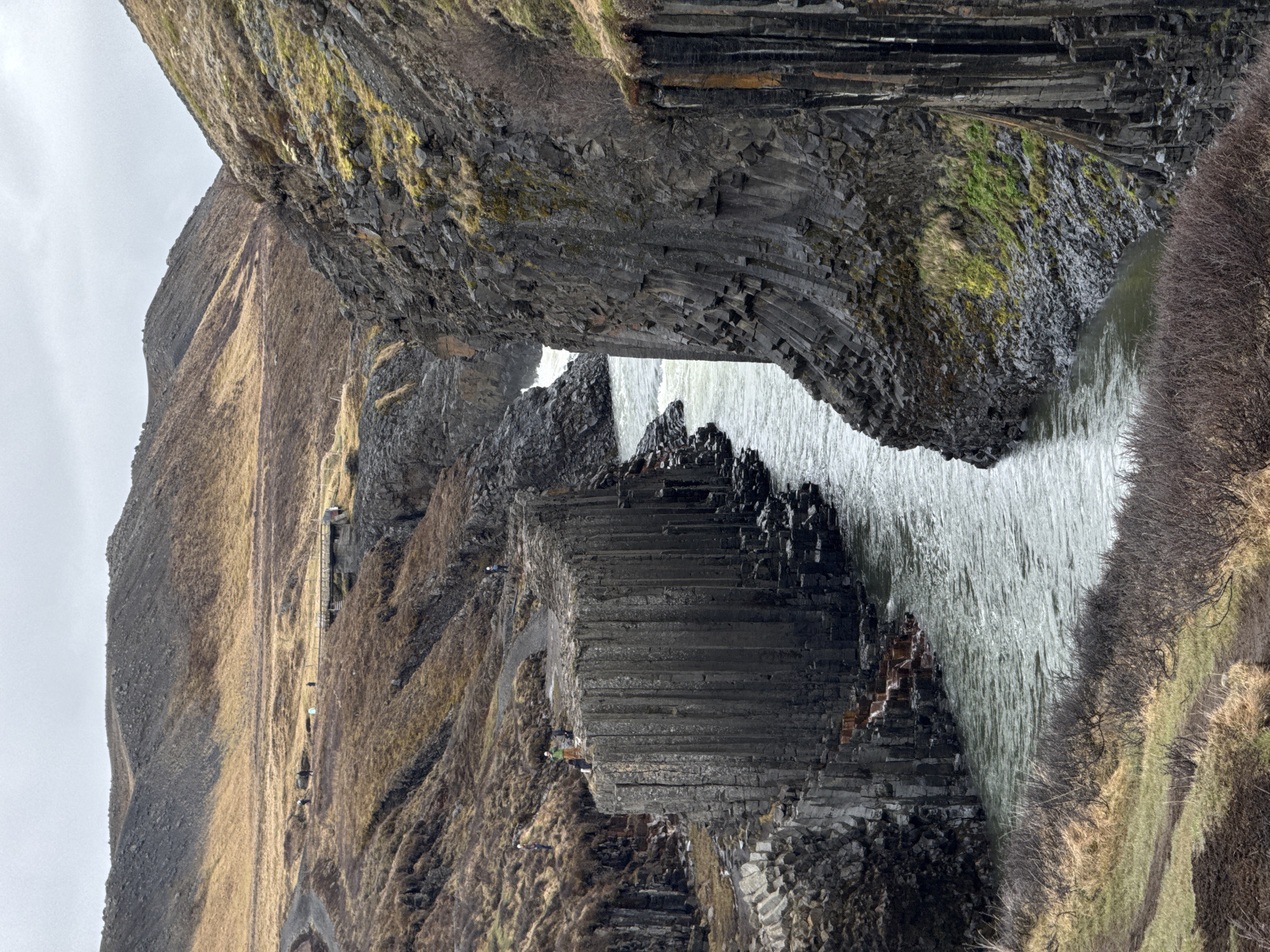 This photograph depicts a powerful waterfall cascading over dramatic columnar basalt formations in a rugged, natural landscape. The scene showcases a wide curtain of white water flowing from an elevated rock structure, potentially through a natural archway, into a river or pool below.
The dominant geological features are the dark, intricate columnar basalt cliffs, which display distinct hexagonal or polygonal patterns. These formations frame the waterfall and line the riverbanks. The surrounding terrain consists of steep, rocky slopes and hillsides, sparsely covered with dry, brownish-green grass and patches of moss, suggesting a temperate or cold climate. Some leafless bushes are visible on the right bank.
Several small human figures are scattered throughout the scene, providing a sense of scale. Multiple individuals are visible walking along a path or trail that traverses the upper left cliff face, where a fence and what appears to be a small structure are also present. Closer to the waterfall, one person in a light-colored top stands on a flat basalt outcrop to the left of the falling water, and another individual wearing a green jacket is situated on a lower basalt section near the river's edge.
The sky is overcast, indicating a daytime scene with diffused light rather than direct sunlight. The overall color palette is dominated by the dark grays of the basalt, the bright white of the cascading water, and muted earthy tones of the sparse vegetation. No visible text is present in the image. The unique geological features strongly suggest a location such as Iceland.