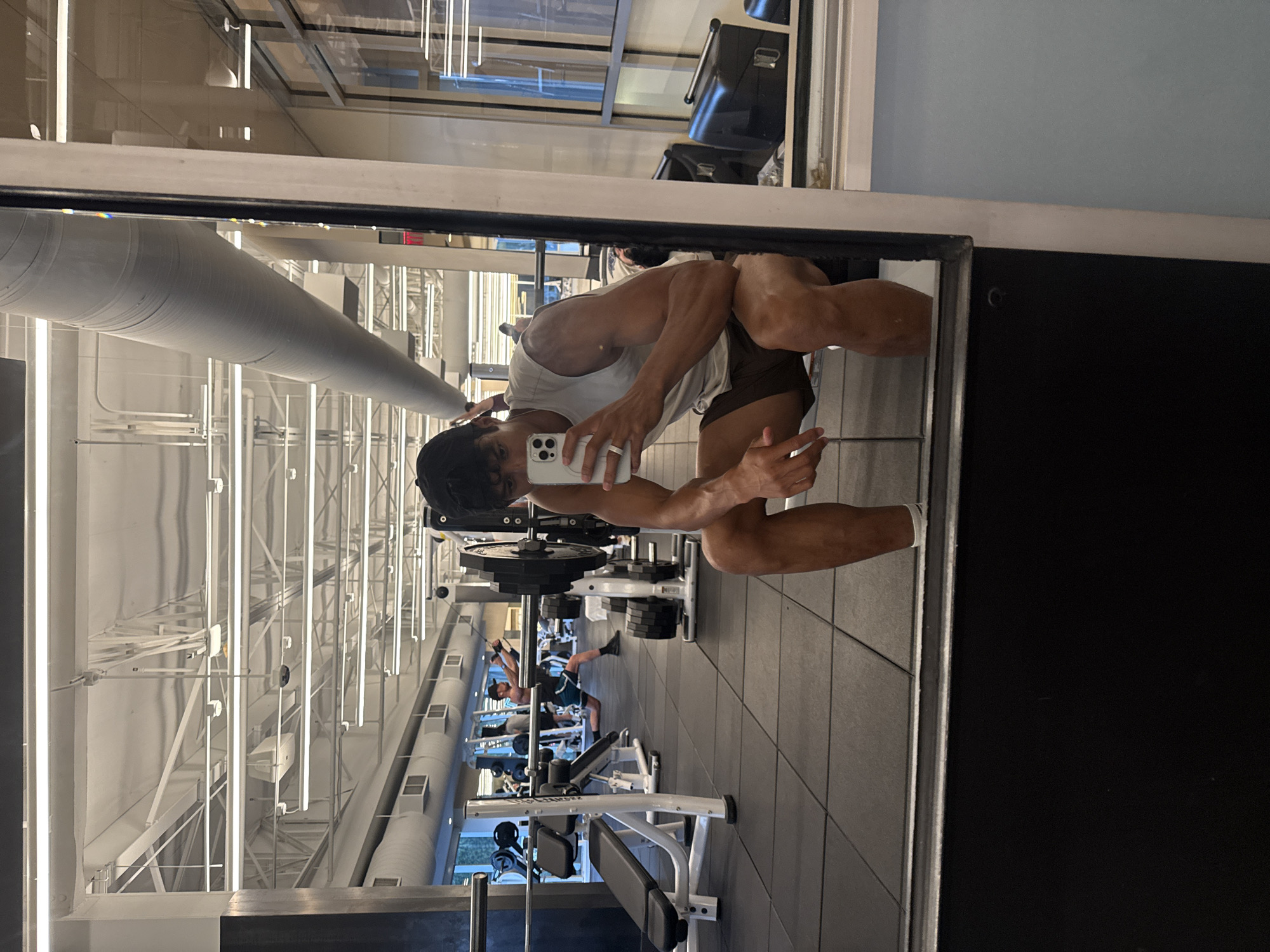 The image is a mirrored selfie of a muscular man in a gym setting. He is wearing a white tank top and brown shorts. He is holding a smartphone in his left hand, presumably taking the picture. Behind him, there are various gym equipments and other people exercising. The gym features a high ceiling with visible pipes and lighting fixtures. The presence of large windows suggests the location is likely in a commercial building. Without external cues it is difficult to pinpoint the exact gym location in West Hollywood.