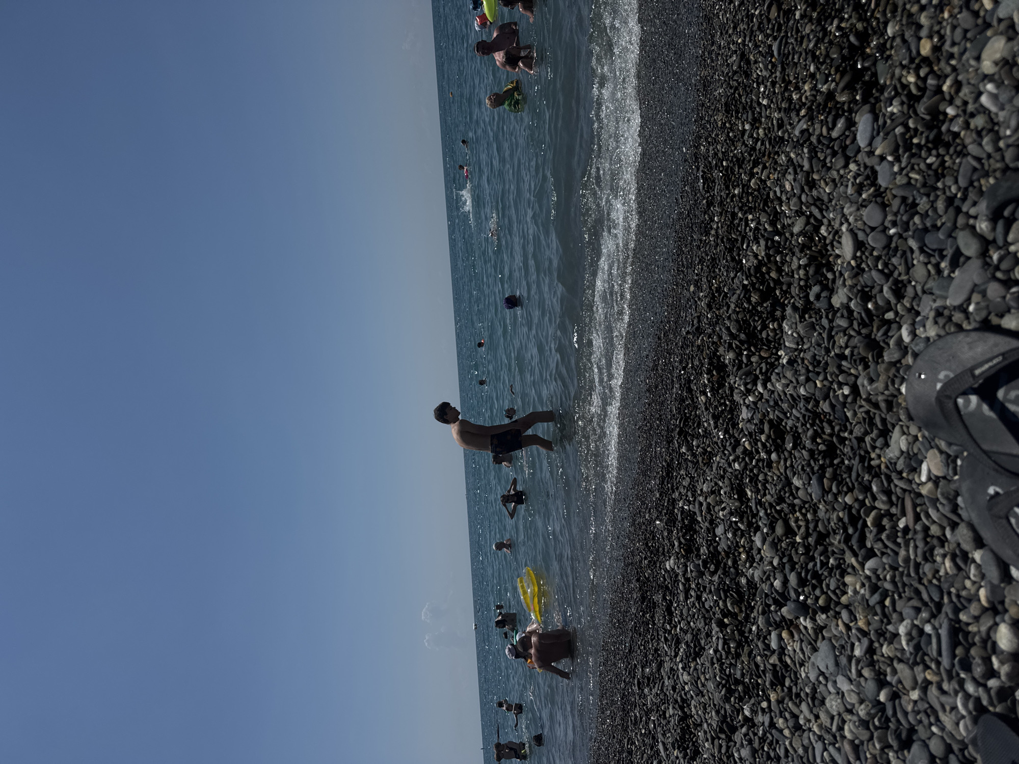 On a clear day with a bright blue sky, people are enjoying the water at a pebble beach in Sochi, Russia. The foreground is dominated by a dark pebble beach, with a few pairs of black flip-flops visible at the bottom. The sea, a deep blue, is populated with numerous individuals swimming and wading. Some are playing with inflatable toys, including a yellow float. A young man in dark swim trunks stands in the shallow water, looking towards the left. Further out, several other swimmers are scattered across the expanse of the sea, with a few small waves breaking on the shore. The horizon is visible, meeting the sky with a slight haze.