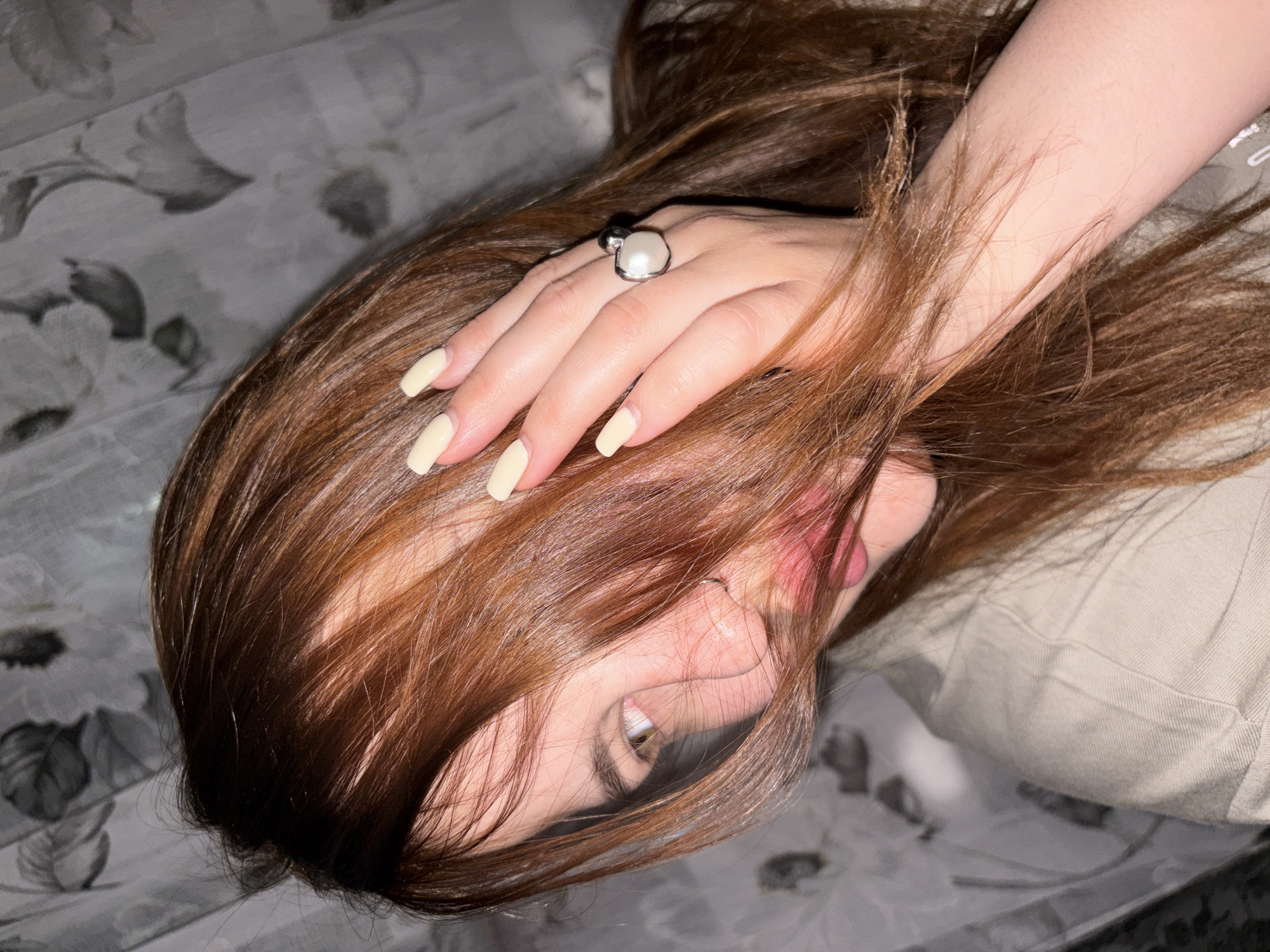 A close-up, high-angle portrait captures a person, likely female, lying down indoors. The subject's head is tilted, with long, straight, reddish-brown hair flowing around and partially covering the face. A hand with long, square-tipped, cream-colored nails is placed over the hair and part of the face, drawing attention to a distinctive silver-colored ring featuring a large, oval, light-colored stone, possibly a pearl.
Only one eye, a segment of the nose, and a portion of the mouth are visible, peeking through the cascade of hair. The visible eye looks towards the viewer, and the lips are slightly parted, suggesting a relaxed or subtly playful expression. The person is dressed in light-colored, possibly beige or tan, clothing, visible on the right side of the frame.
The background consists of a muted, monochrome floral patterned fabric, likely bedding or upholstery, in shades of grey and white. The lighting appears soft and indirect, indicating an indoor setting. The image conveys a calm and intimate mood, without any visible text. The general location context for this media is Port-Katon, Russia.