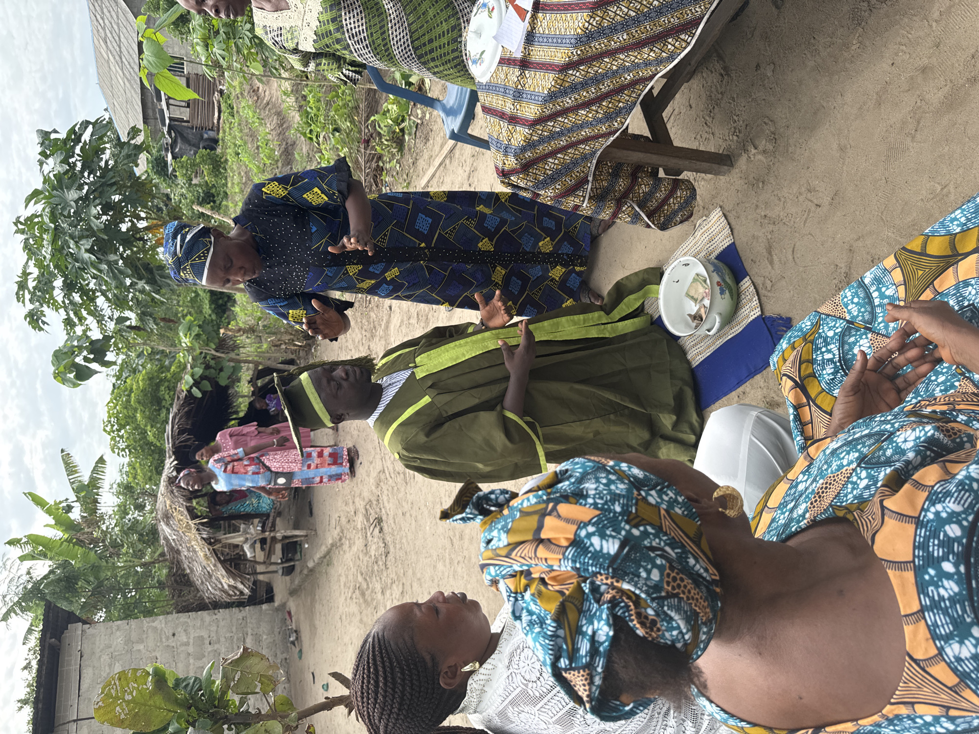 A gathering of people is occurring outdoors in Tofa, Nigeria. A woman in a blue and yellow patterned dress stands, gesturing with her hands as she speaks to a man in a green robe and hat seated on the ground. A cup with money is beside him. Other women, some wearing patterned headscarves and dresses, are seated around them. A table covered with a patterned cloth is to the right of the standing woman. A simple structure with a thatched roof is visible in the background among trees and a cinder block building. The ground is sandy.
