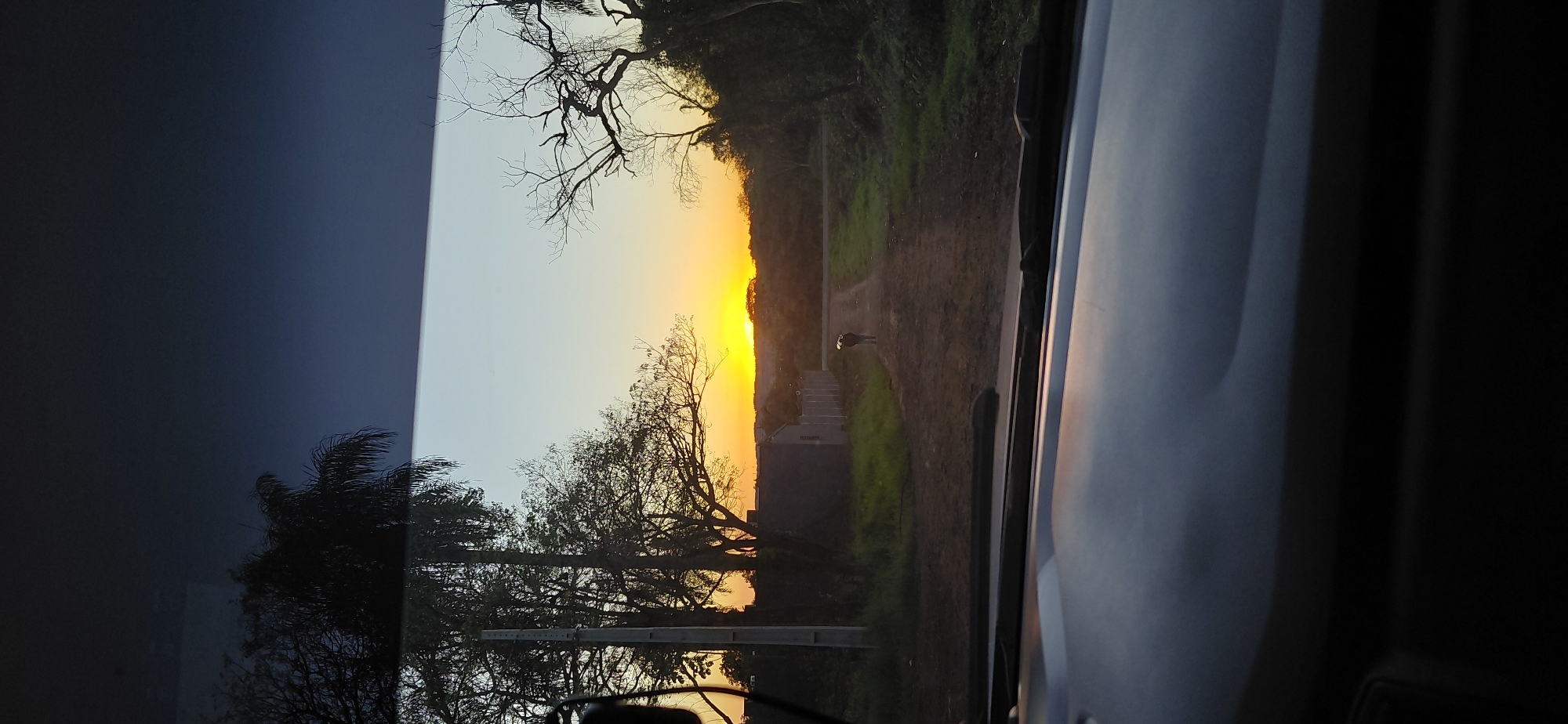 This image captures a sunset viewed from inside a vehicle, likely on the outskirts of Encarnación, Paraguay. The perspective is from the driver's seat, looking through the windshield.
In the foreground, the dark, textured dashboard of the car is visible, reflecting the sky. Beyond the dashboard, a dirt road stretches into the distance, lined by sparse vegetation and some low-lying buildings with fences. The sky is a gradient of pale blue at the top, transitioning to a vibrant orange and yellow near the horizon, indicating the setting sun. Silhouettes of trees and some foliage frame the landscape.
On the dirt road, a lone figure, appearing to be a person walking away from the camera, is visible in the middle ground. The figure is carrying something over their shoulder. The overall atmosphere is serene, with the warm colors of the sunset creating a peaceful ambiance. The setting appears to be rural or semi-rural, possibly on the edge of the city, where the natural landscape meets developing areas.