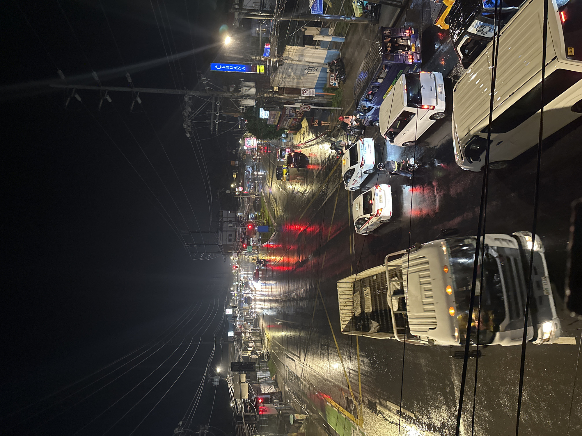 This is a nighttime, high-angle, medium shot of a wet street in Davao City, Philippines, during what appears to be a rainstorm. The road is heavily reflecting the lights of traffic signals, streetlights, and vehicle headlights, creating a shimmering effect. Various vehicles, including a white flatbed truck in the foreground, white sedans, a jeepney, and motorcycles, are visible on the road, some with their lights on. Traffic signals are illuminated red. Overhead, a dense network of power lines and utility poles crisscrosses the sky. Buildings line both sides of the street, some with illuminated signage, including a prominent blue sign that reads "CEBUANA." The overall atmosphere is dark and moody, with the rain and reflections creating a dramatic urban scene.