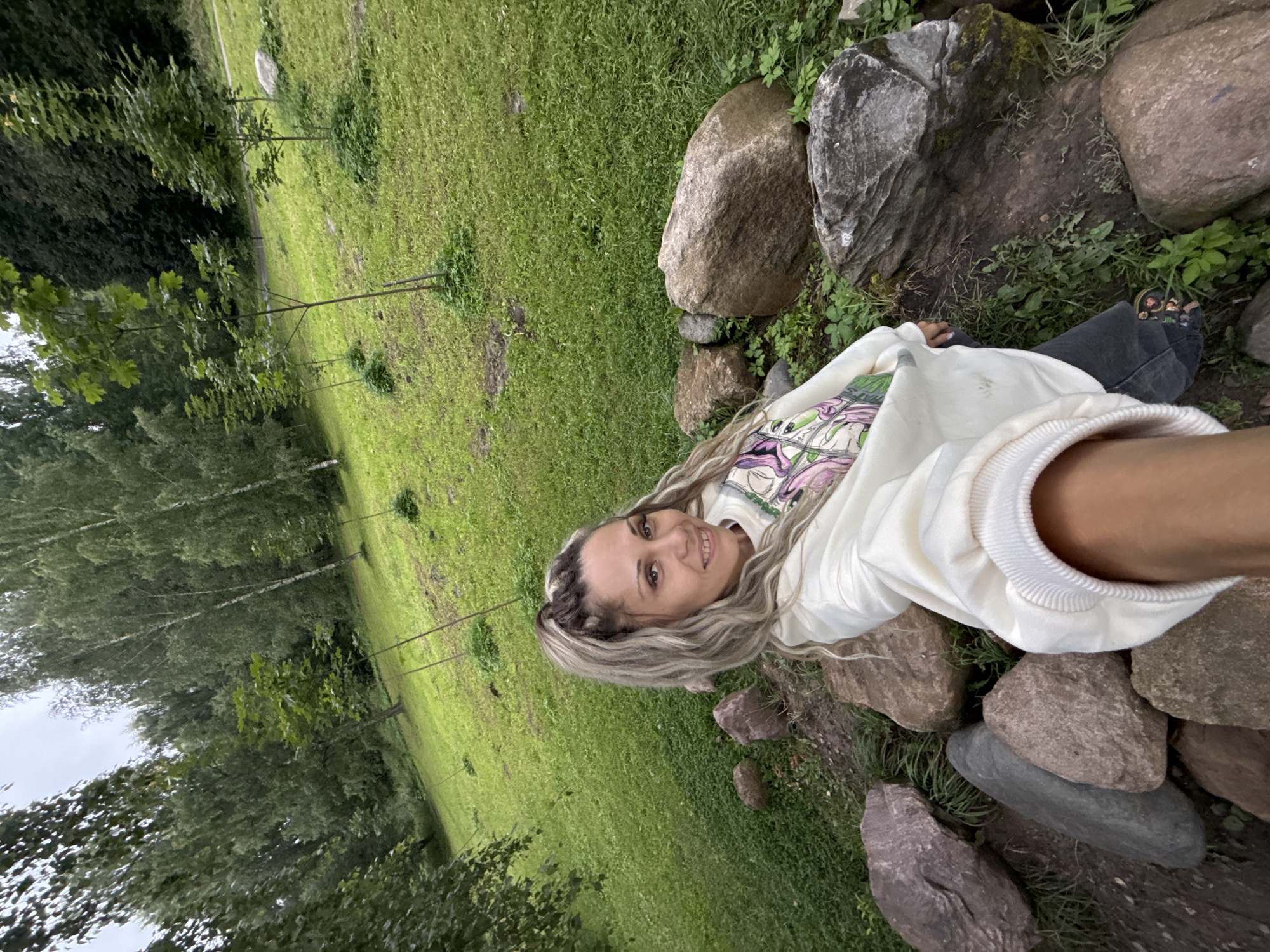 A woman with blonde dreadlocks, wearing a white sweatshirt with a graphic print and dark pants, is taking a selfie in a park-like setting. She is positioned in the foreground, with large rocks around her feet. The background consists of a grassy slope dotted with young trees and mature trees. The sky is overcast. The image appears to be taken in Rusanovo, Russia, given the information provided.